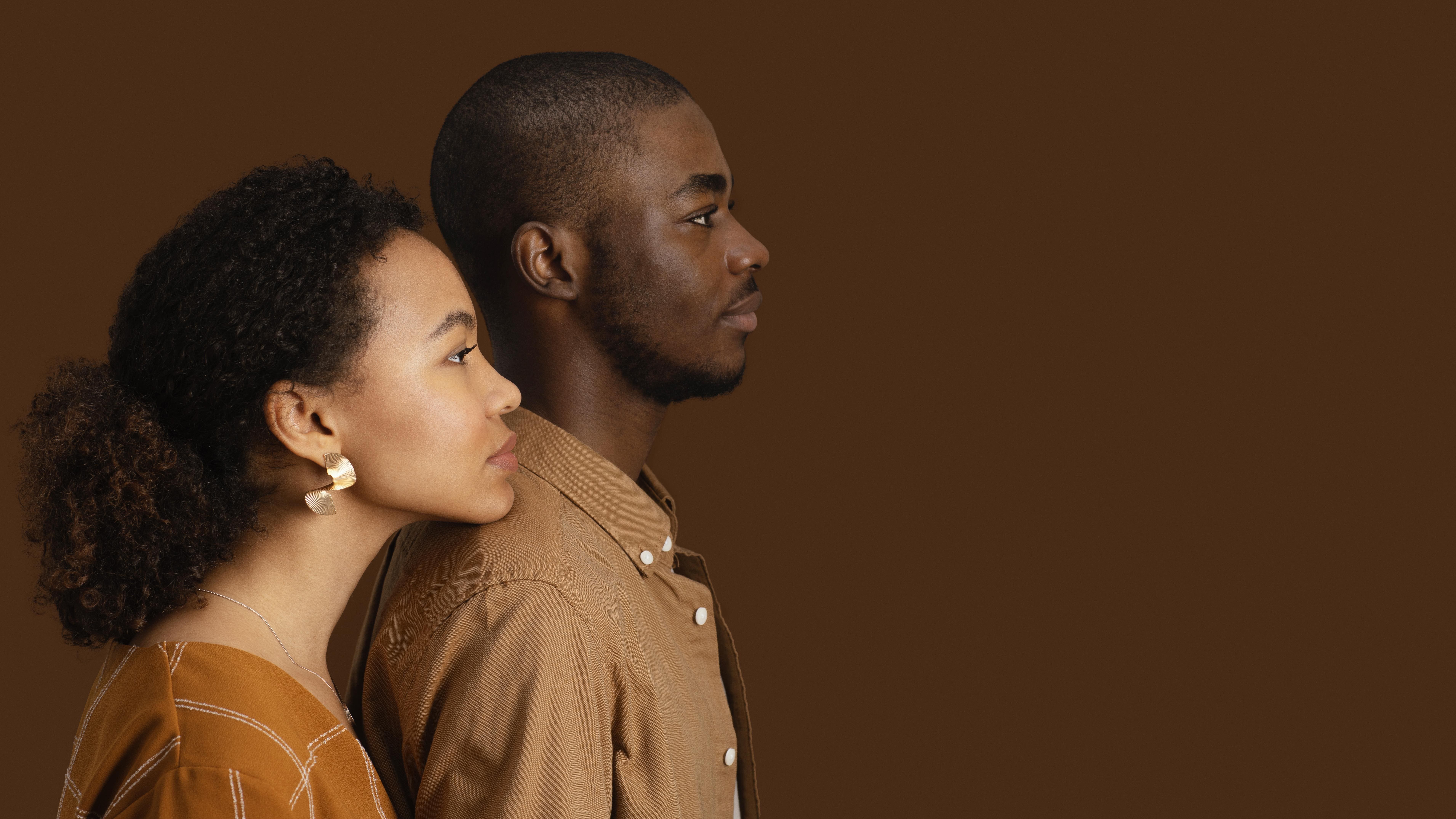 A imagem mostra um casal em perfil, ambos olhando para frente, contra um fundo marrom sólido. A mulher, à esquerda, tem cabelo cacheado preso e usa brincos dourados grandes. Ela veste uma blusa em tons terrosos. O homem, à direita, tem cabelo curto, barba rala e também veste uma camisa marrom. Ambos têm uma expressão serena e parecem estar lado a lado, transmitindo uma sensação de união e tranquilidade.