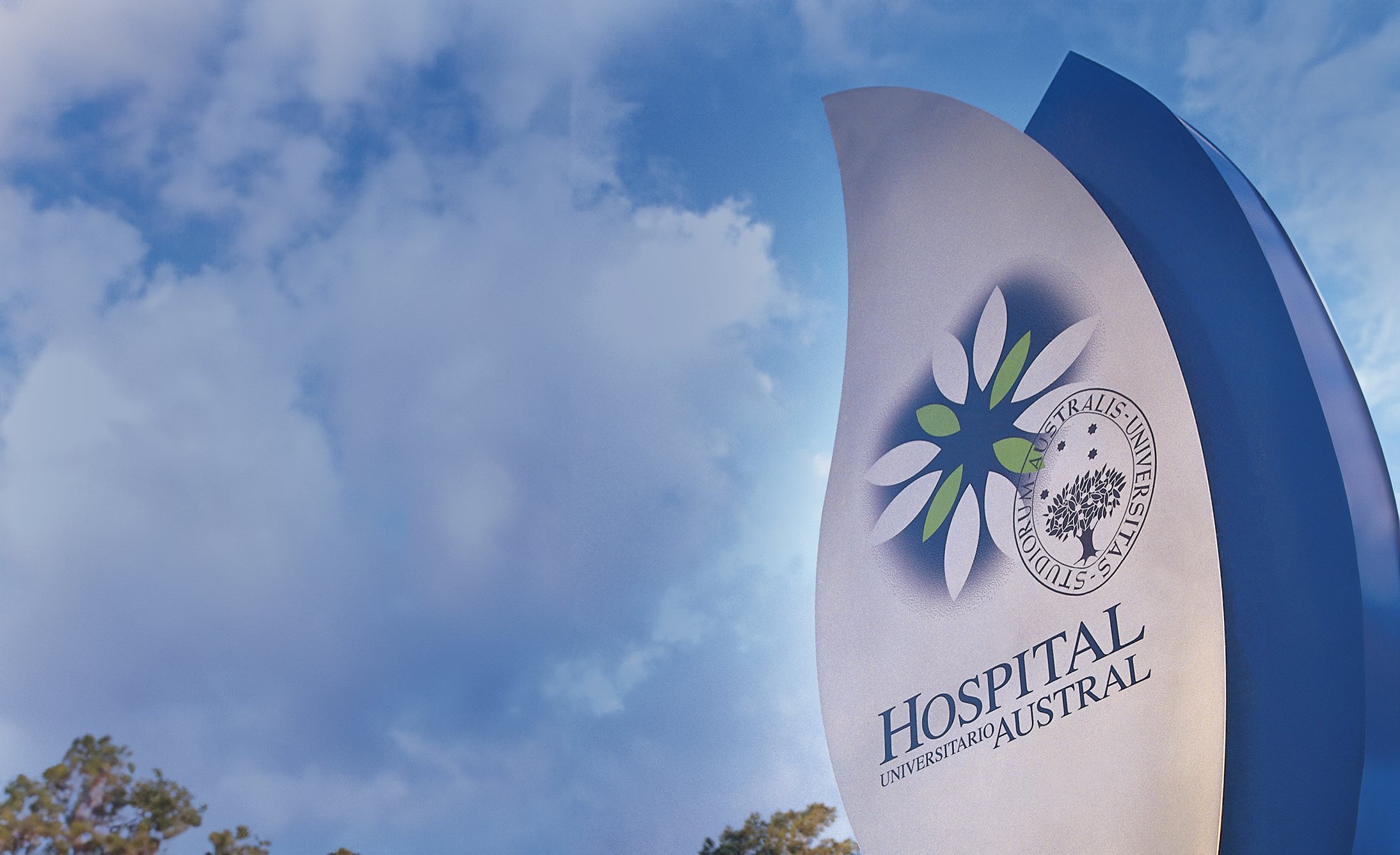 Sign for Hospital Universitario Austral against a partly cloudy sky. The sign features a circular emblem with leaves and a tree, conveying a serene atmosphere.