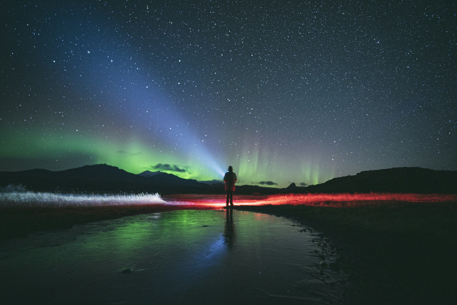 Person standing beneath the northern lights in Iceland with mountains and a star-filled sky in the background.