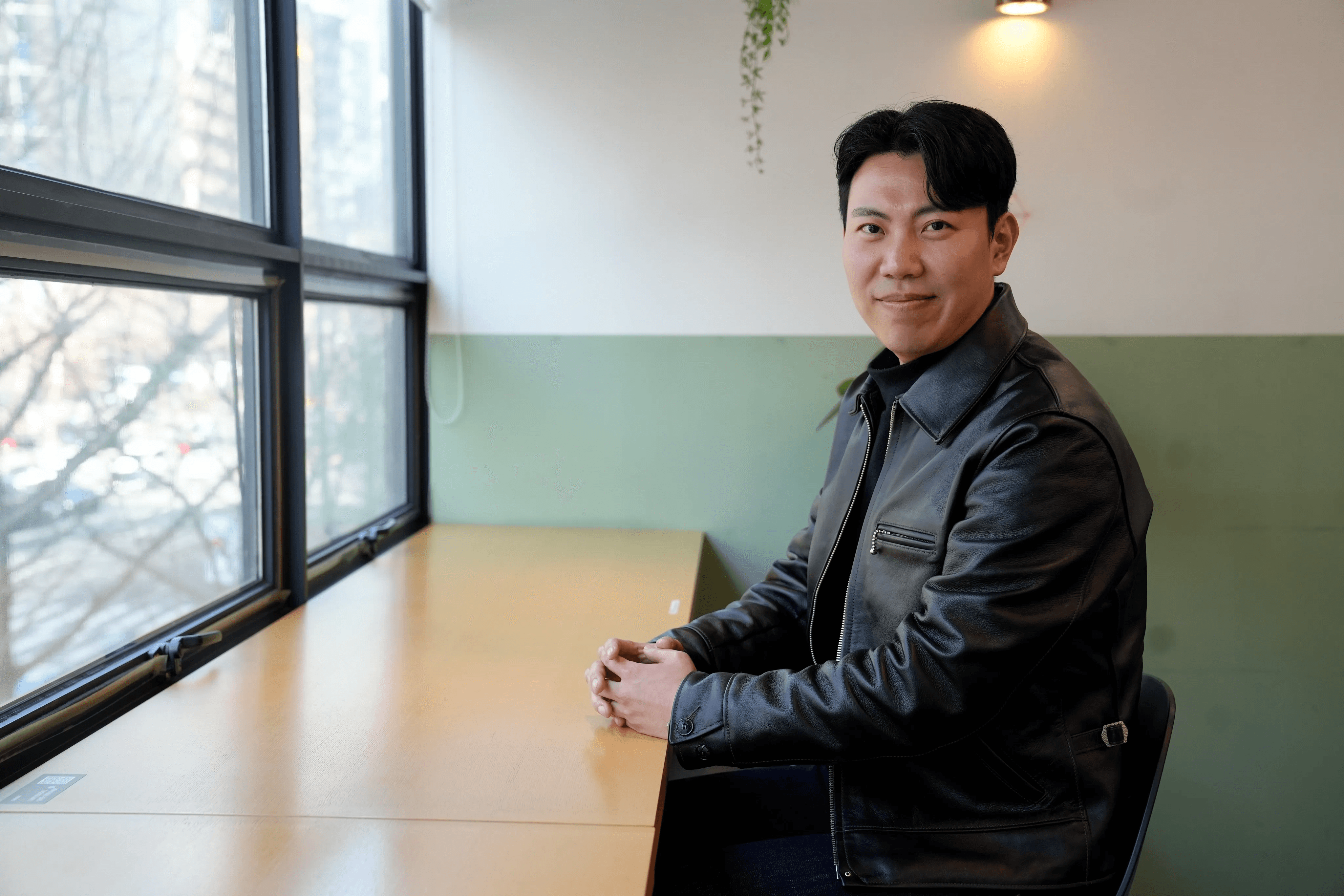 창가 옆 테이블에 앉아 정면을 바라보며 미소 짓고 있는 남성. 검정 가죽 재킷 착용. 배경에 녹색 벽면과 식물, 외부 겨울 나무가 보임.