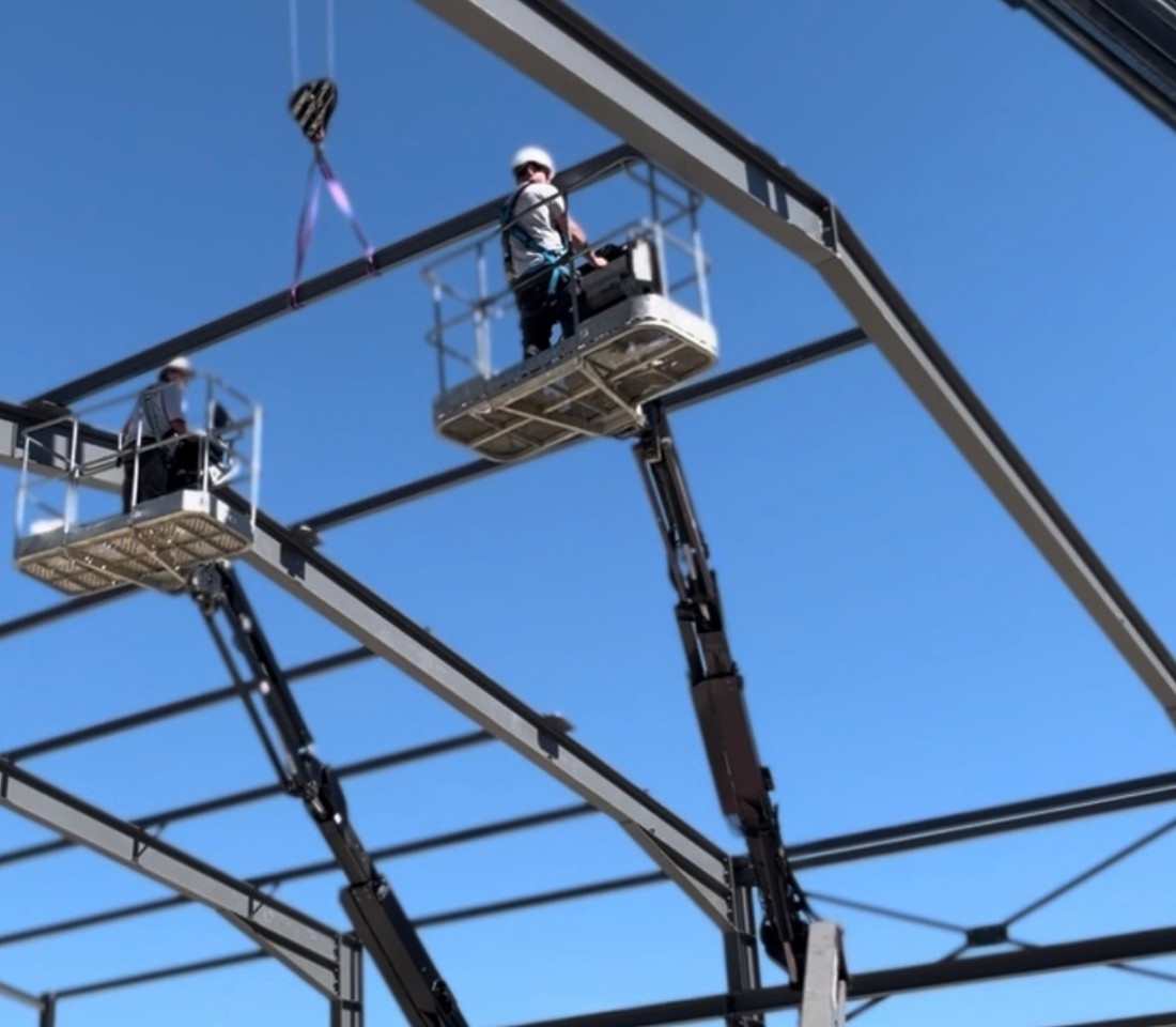 Montage de la structure porteuse d’un bâtiment industriel en acier par deux techniciens sur nacelles hydrauliques.
