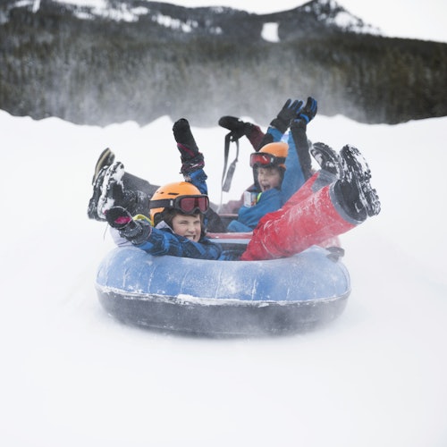 Three children in helmets ride an inflatable snow tube down a snowy slope, smiling with arms and legs raised.