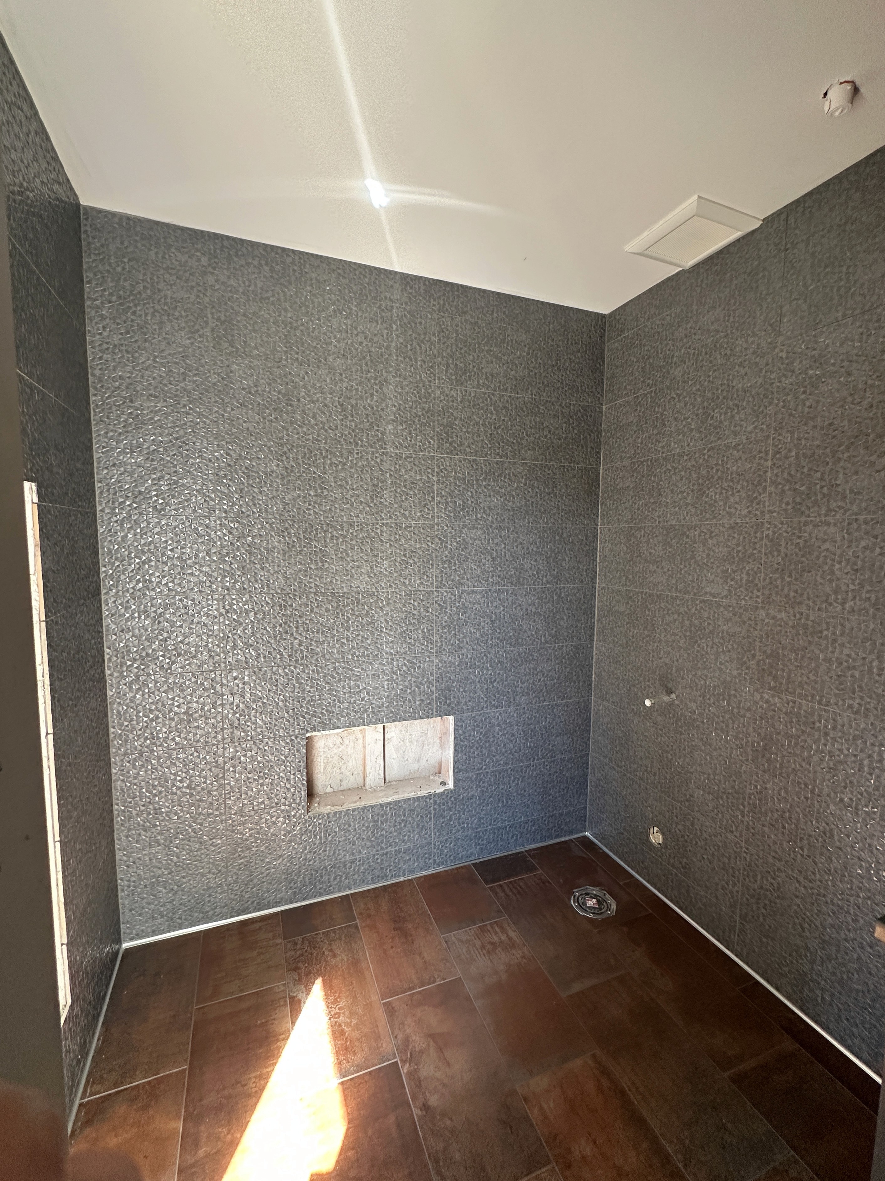 Contemporary bathroom with dark textured wall tiles and wood-look floor tiles, showcasing a built-in wall niche and plumbing setup.