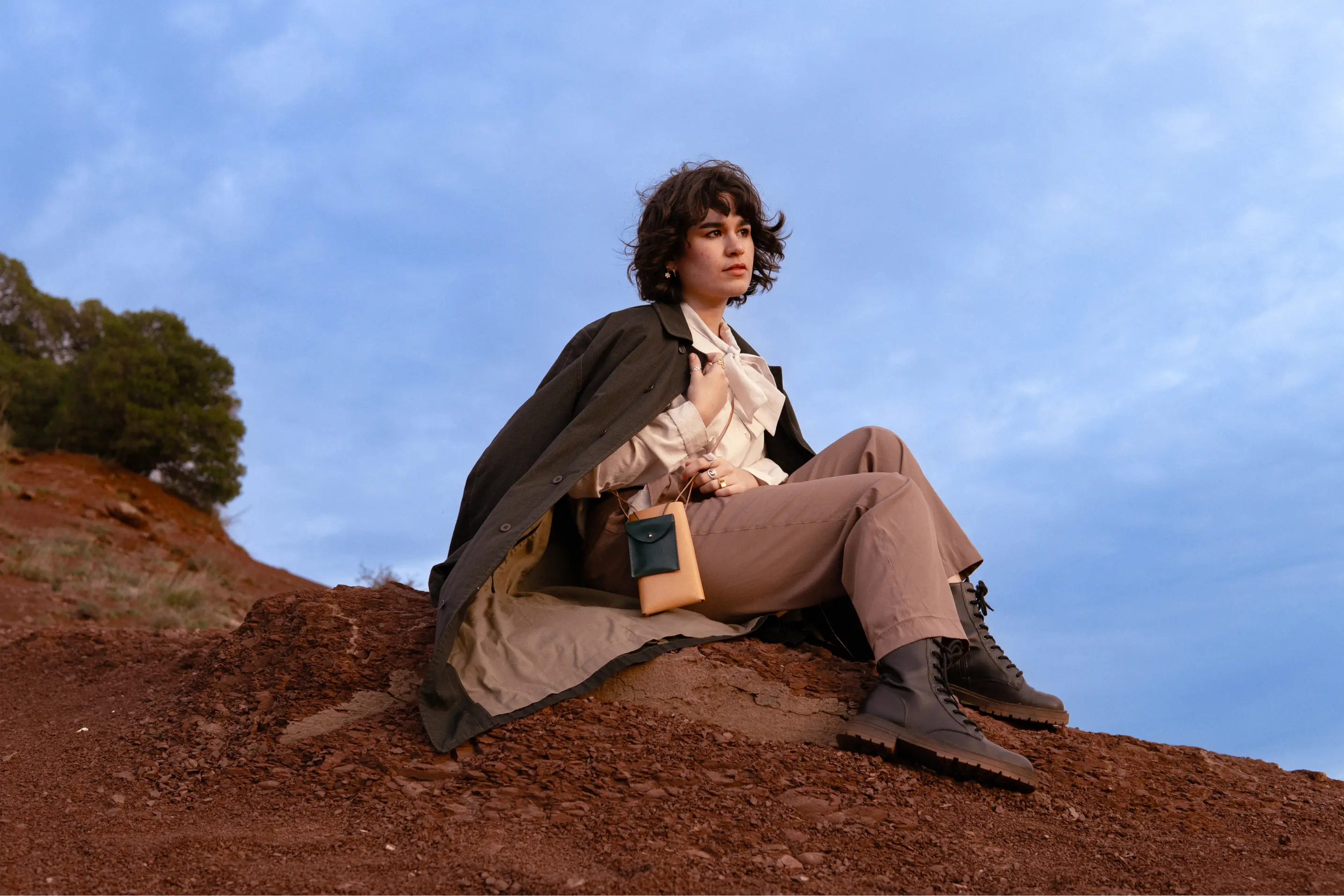 Anaïs Brancheriau standing on ochre-colored ground, wearing a long dark coat and a leather pouch under a blue sky, with a second blurred figure in the foreground.