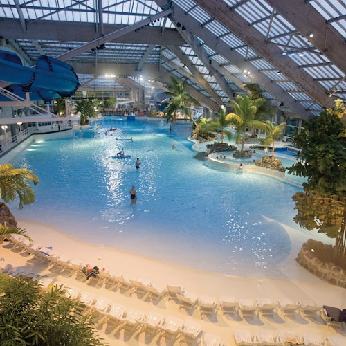 Indoor water park with a large pool, palm trees, lounge chairs, and a blue water slide. Several people are in the water.