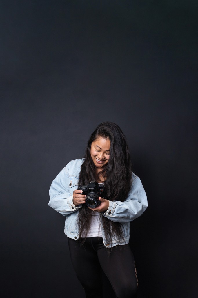 Photographer in a casual denim coat posing with her camera on a dark blue background in a Winnipeg studio