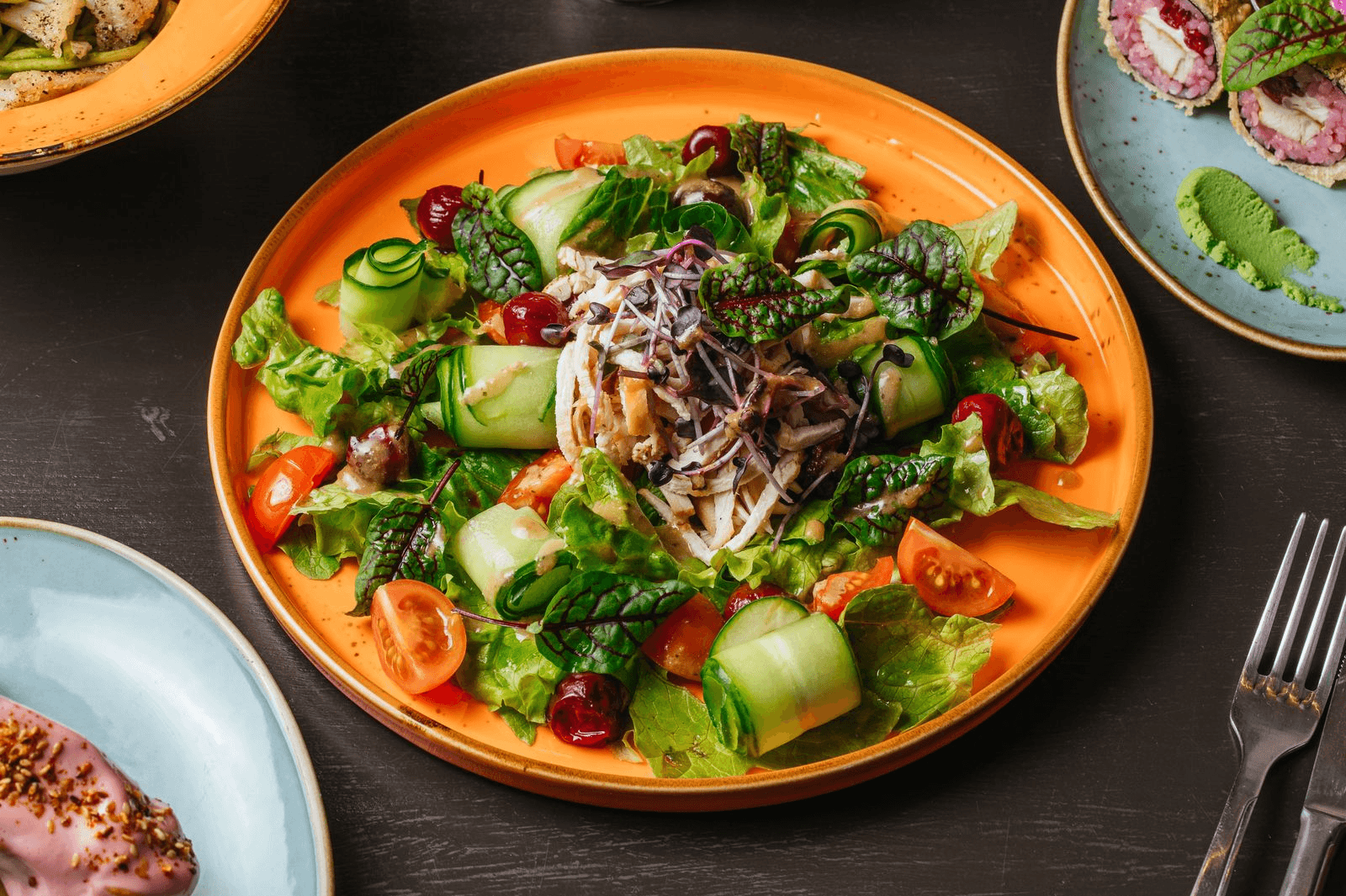 A vibrant salad on an orange plate features mixed greens, cucumber rolls, cherry tomatoes, and shredded radish. Nearby, a fork and other colorful dishes add to the fresh, appetizing scene.