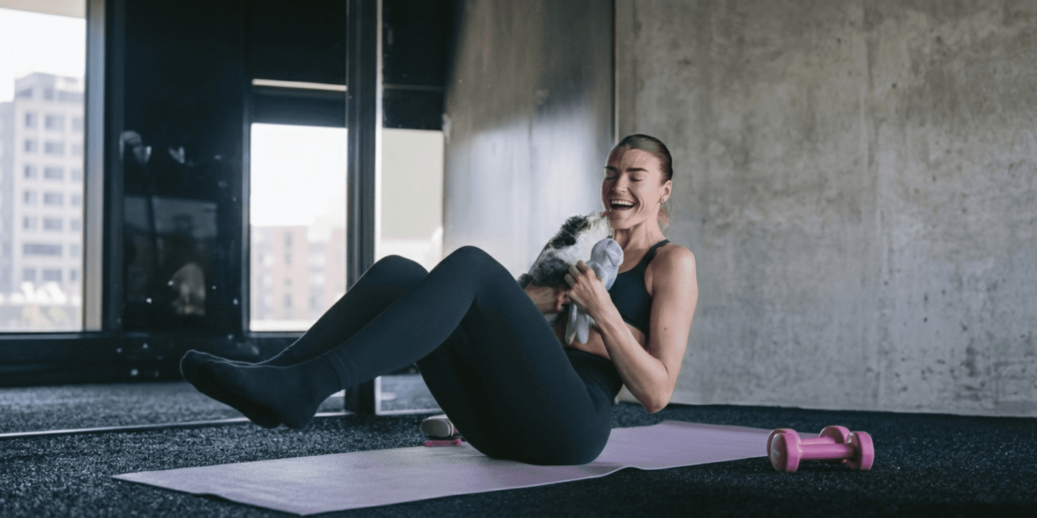 Woman doing Pilates on a mat while smiling and holding a playful puppy indoors.