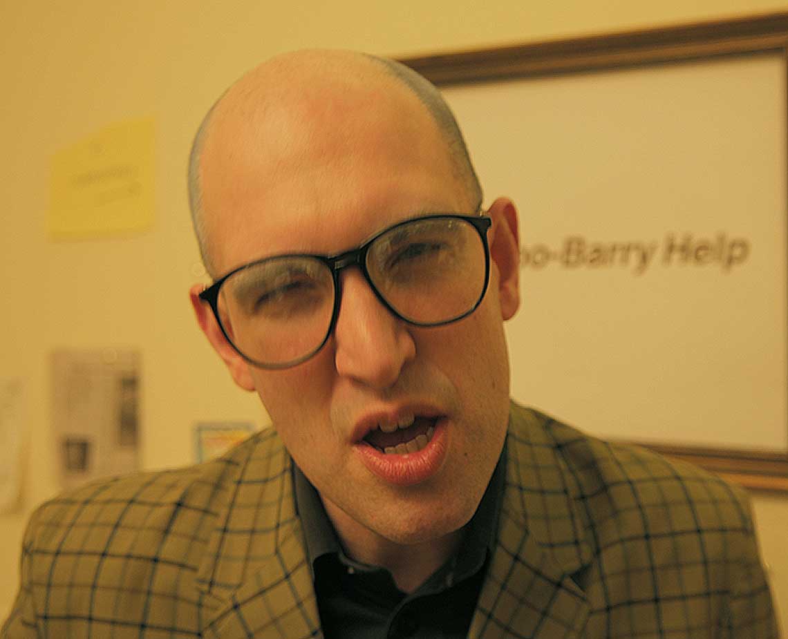 The image shows a man with a shaved head, wearing black-framed glasses and a light olive green or khaki plaid blazer over a dark-colored collared shirt. He is looking directly at the camera with his mouth slightly open. The background features a light beige or cream-colored wall with a framed sign that reads "Barry Help" in dark text. A portion of a yellow sticky note is visible on the left edge of the image.