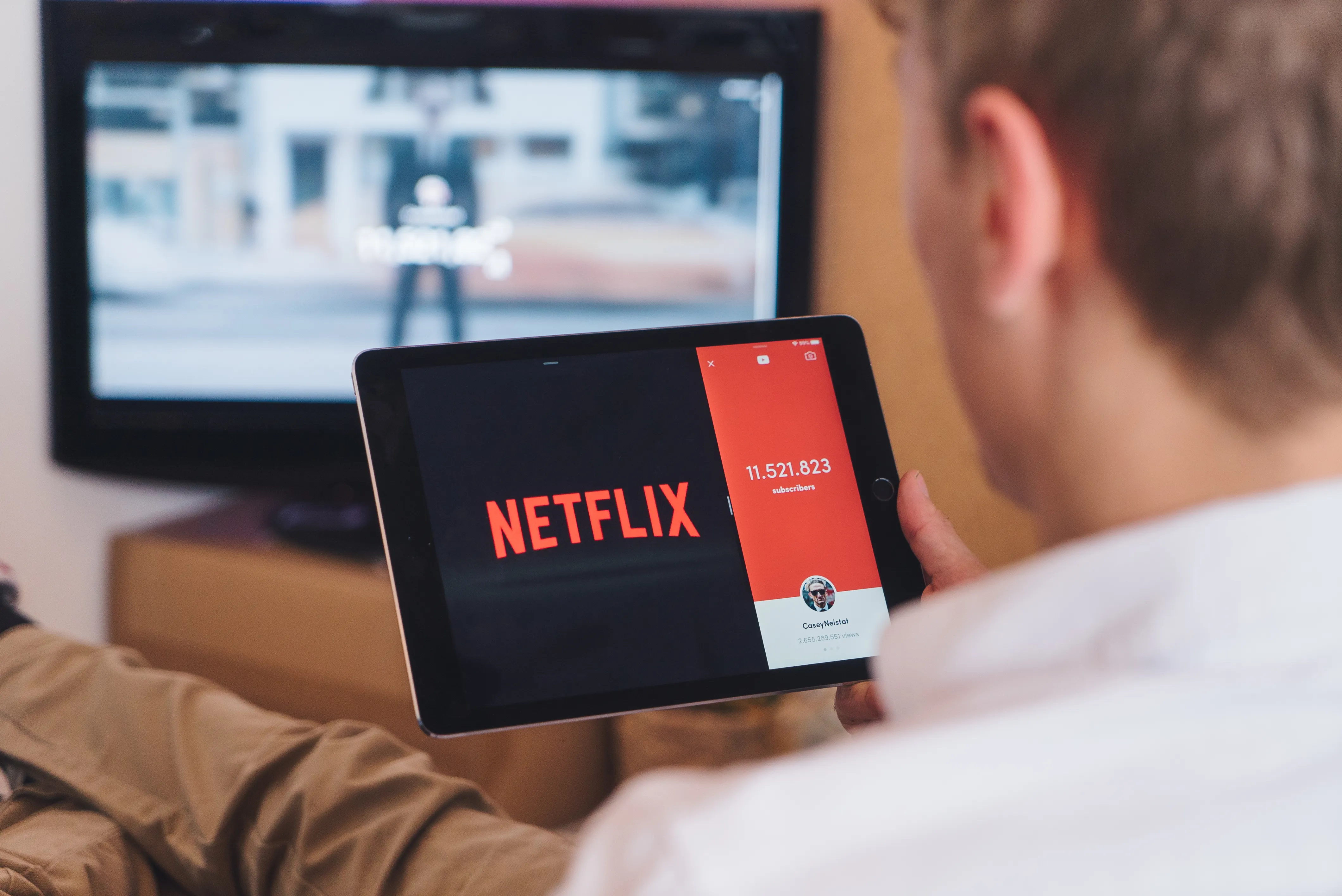 Person holding a tablet displaying the Netflix logo, sitting in front of a television