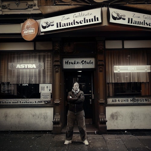 A person stands with arms crossed in front of a dimly lit bar named "Zum Goldenen Handschuh," with signage and curtains in the windows.