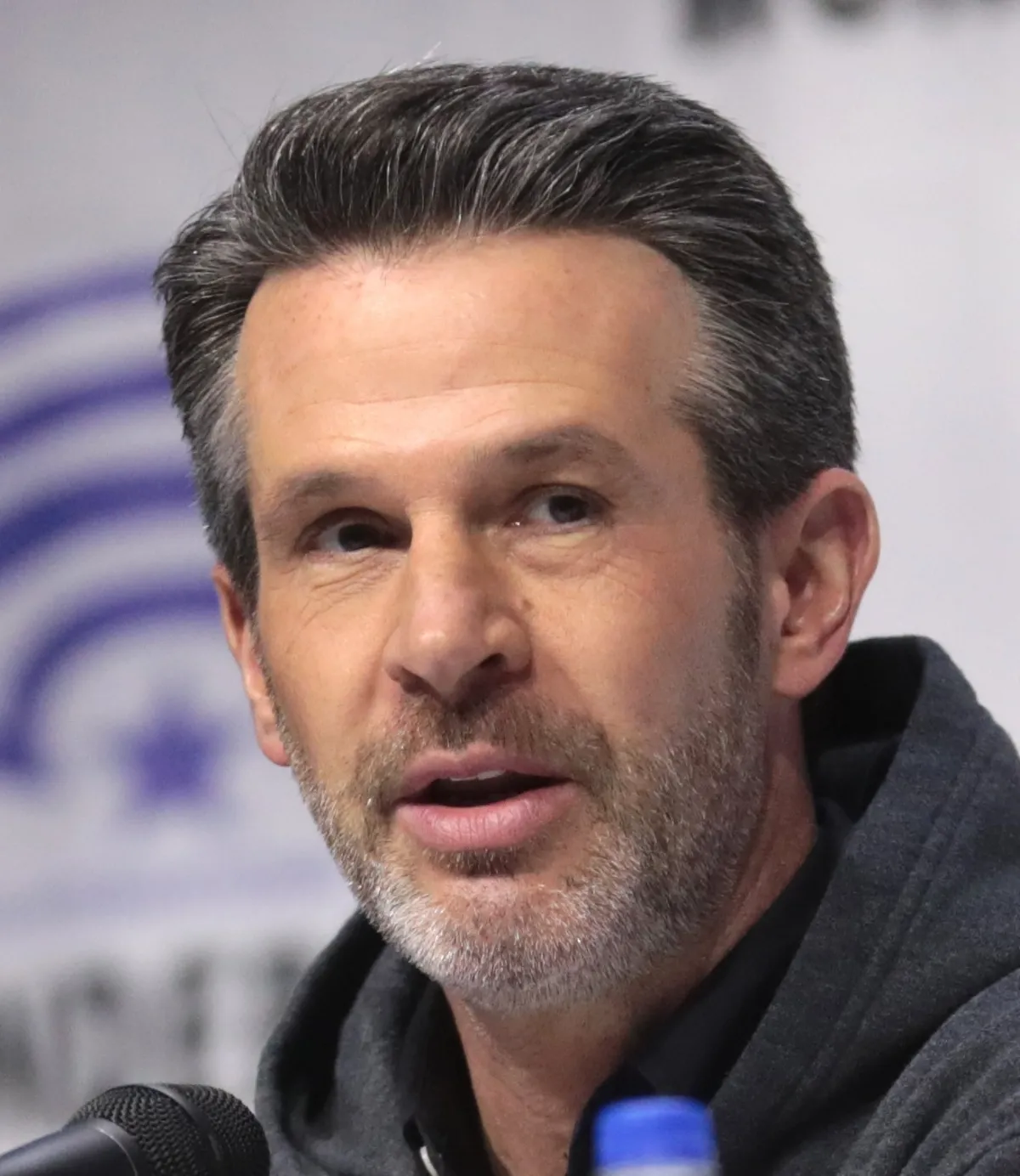 A man with short, brown hair and a greying beard is speaking into a microphone at a press conference, wearing a dark hoodie with a blurred logo in the background.