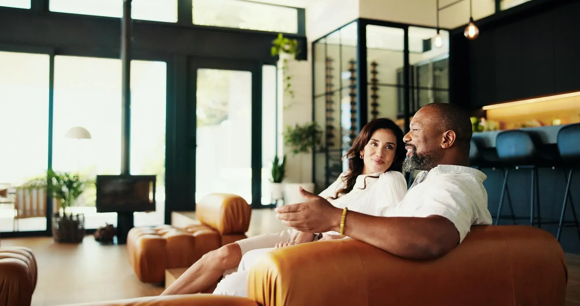 A couple relaxes on a leather sofa in a modern luxury home with floor-to-ceiling windows and elegant decor. The image represents high-value home financing options available through Jumbo Loans from Chris Lewis Home Loans.