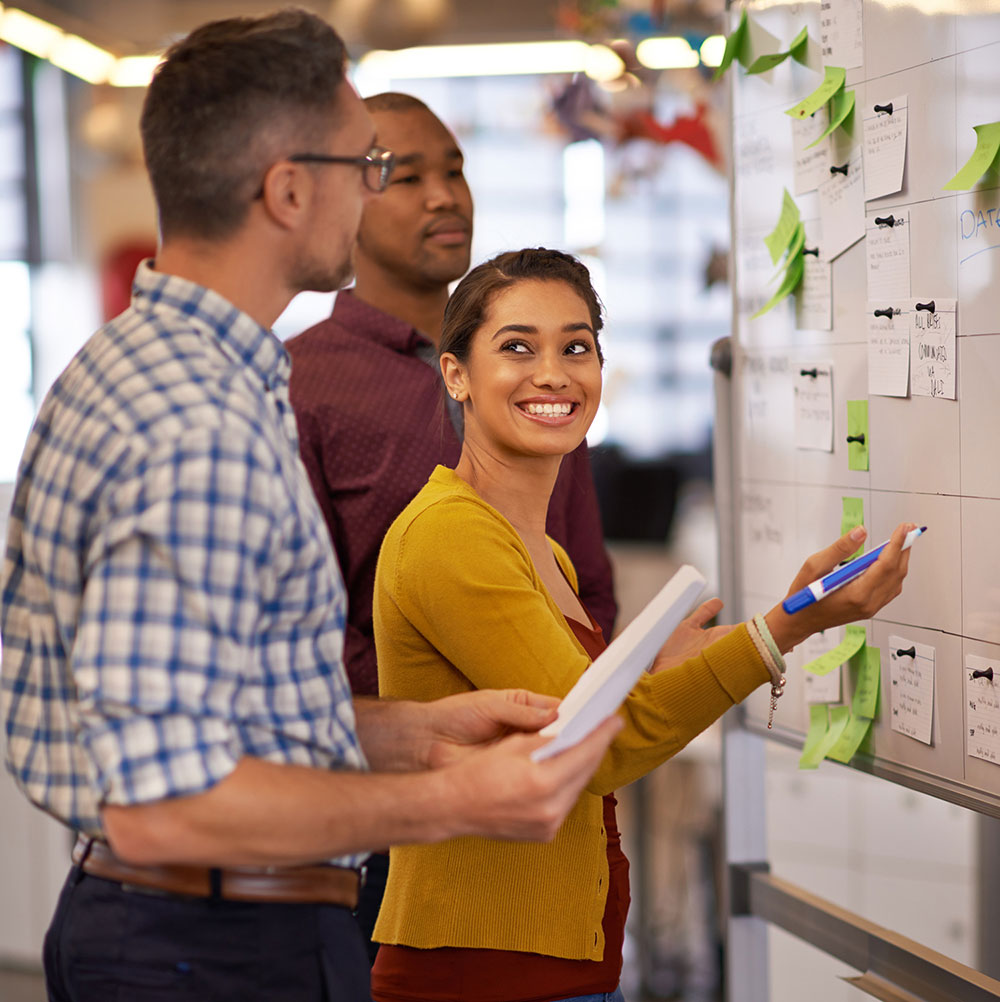 Three colleagues collaborate around a wall-mounted board covered with papers, notes, and green sticky notes held by binder clips. A woman in a mustard yellow cardigan smiles while holding a blue pen and documents, flanked by two male colleagues in casual business attire. The bright, modern office setting suggests an active brainstorming or project planning session.