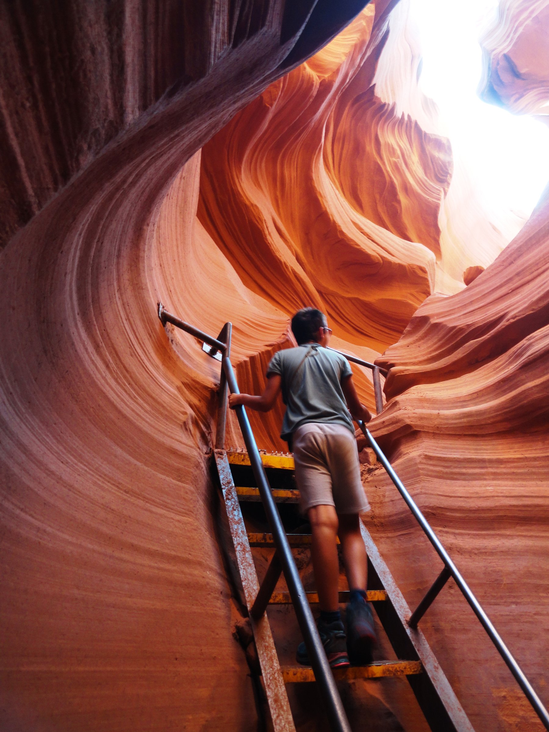 A visitor climbing a steel stairway with handrails inside Lower Antelope Canyon, the narrow orange walls rising on both sides.