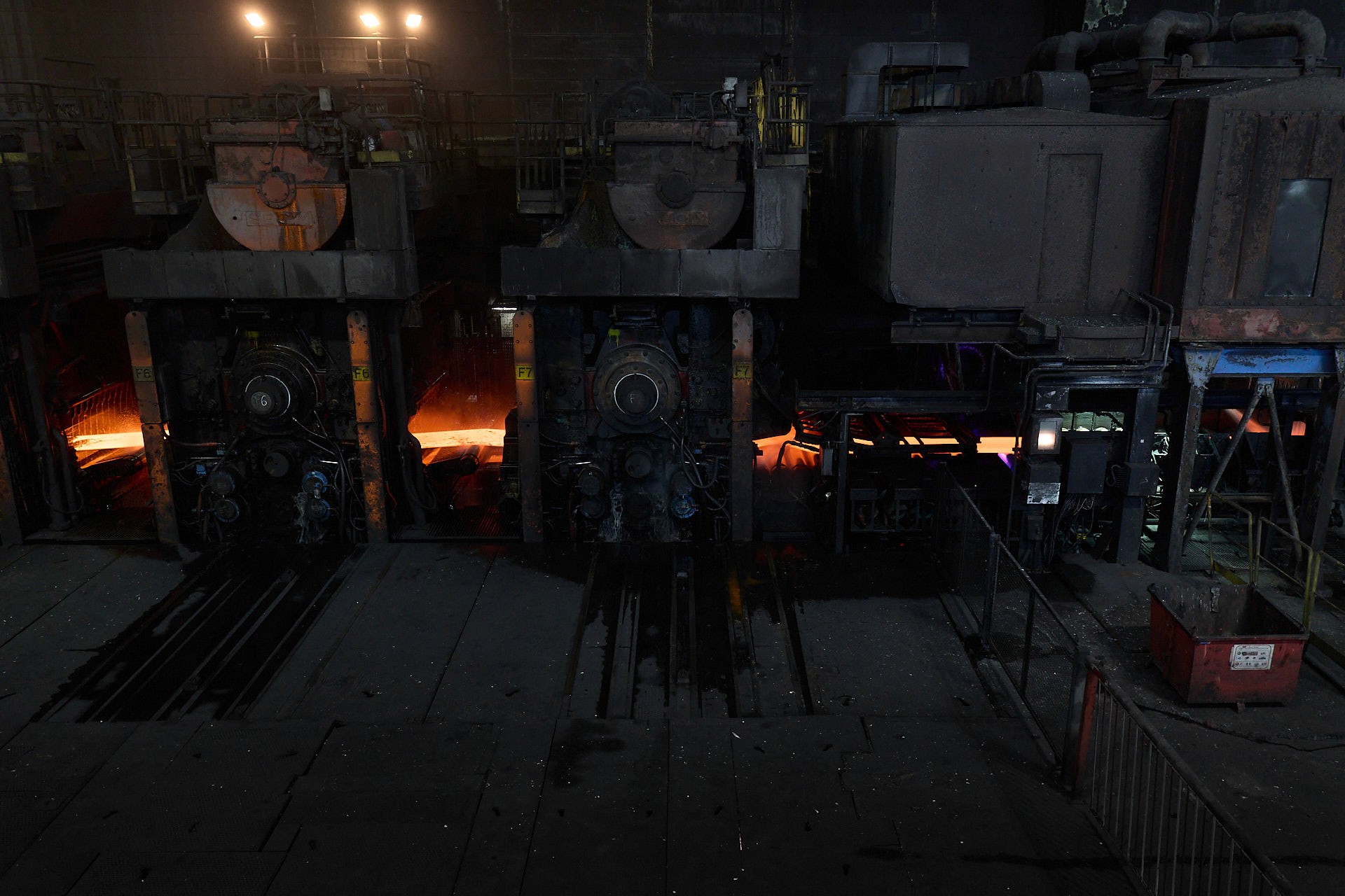 Vue d'ensemble en plongée d'une halle industrielle sidérurgique chez ArcelorMittal à Dunkerque, montrant la ligne de production, la fumée, laminoirs industriel et des brame rouge vif en action.