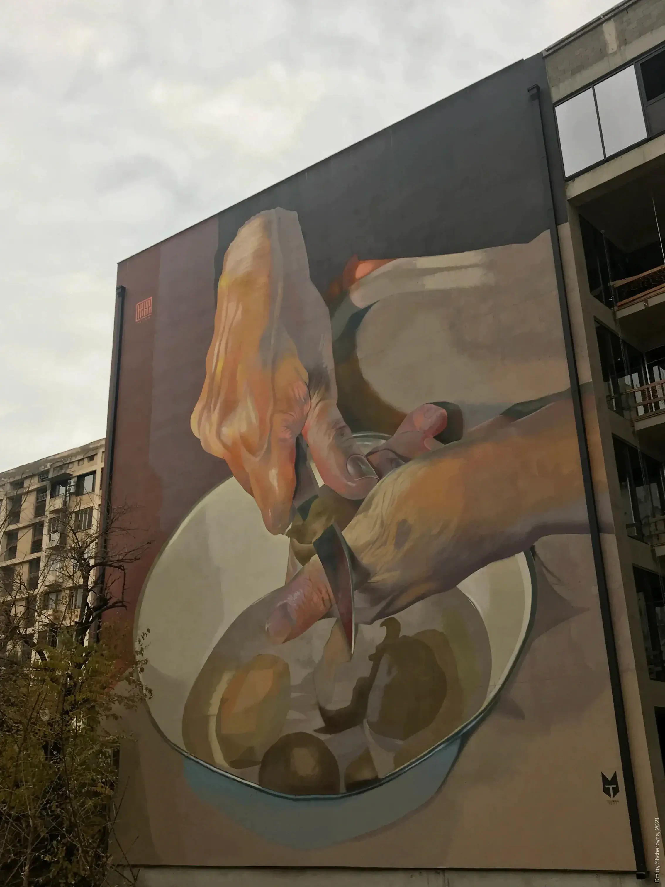 A photorealistic mural on the side of a building, showing a close-up of two hands peeling potatoes with a knife over a bowl, rendered in warm, earthy tones.