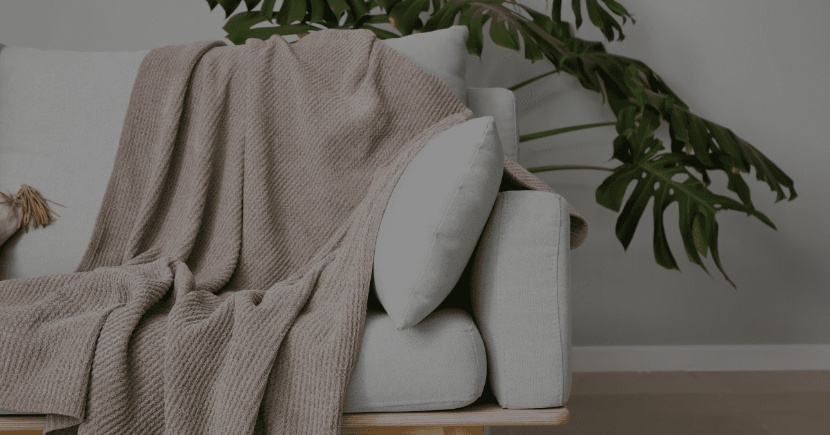 The front view of a grey chair with a warm cozy blanket draped over the edge. A green tropical plant with large leaves sits behind the chair.