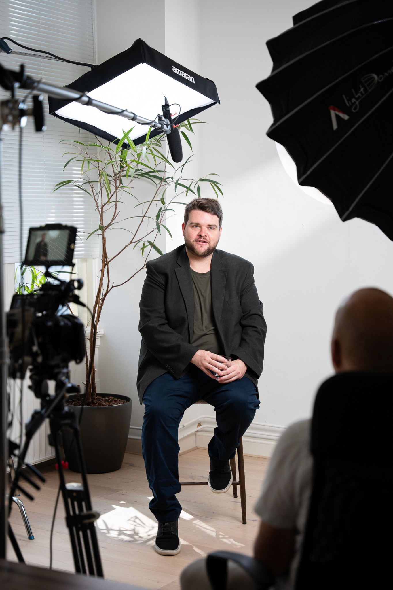 A man in a dark blazer speaking on camera during a video shoot, framed between two cameras and softbox lighting rigs against a white wall with a potted plant behind him.