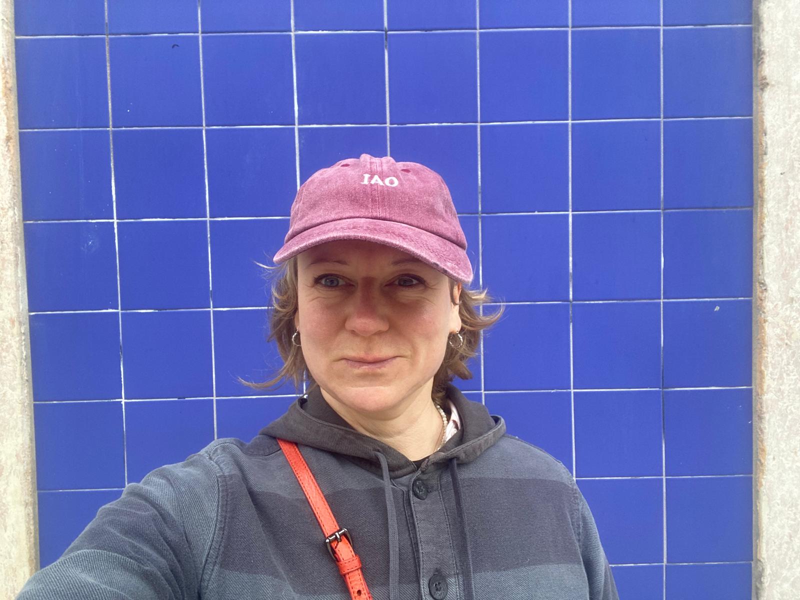 Person wearing a blue puffer jacket and red backpack standing in front of a light blue building on a city street.