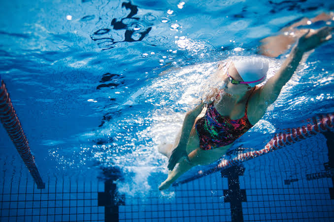 An underwater, wide-angle action photograph of a female swimmer gliding through a bright blue indoor swimming pool. Wearing a white swim cap, colorful patterned swimsuit, and tinted goggles, she reaches forward with one arm while bubbles and turquoise spray ripple around her. The tiled pool floor and lane divider lines are visible in the background beneath shimmering caustic light patterns.