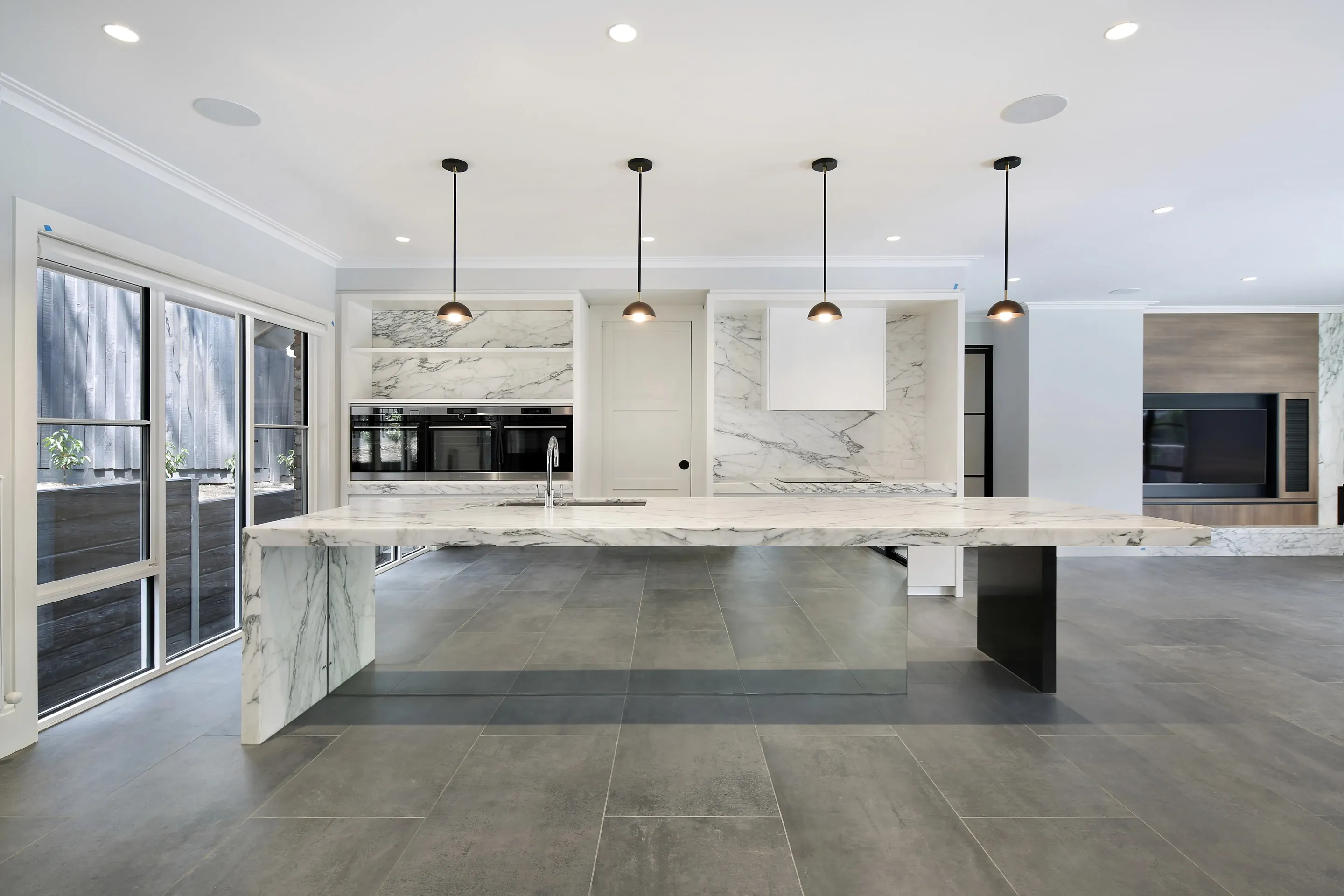 Open-plan kitchen with a large book-matched Calacatta marble island on a black steel leg, copper dome pendants, marble splashback, integrated ovens and charcoal stone tiled floors