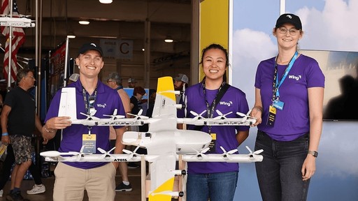 Three people wearing purple shirts stand smiling behind a large, white, drone model with yellow accents, showcased in a bright exhibition space with banners in the background.