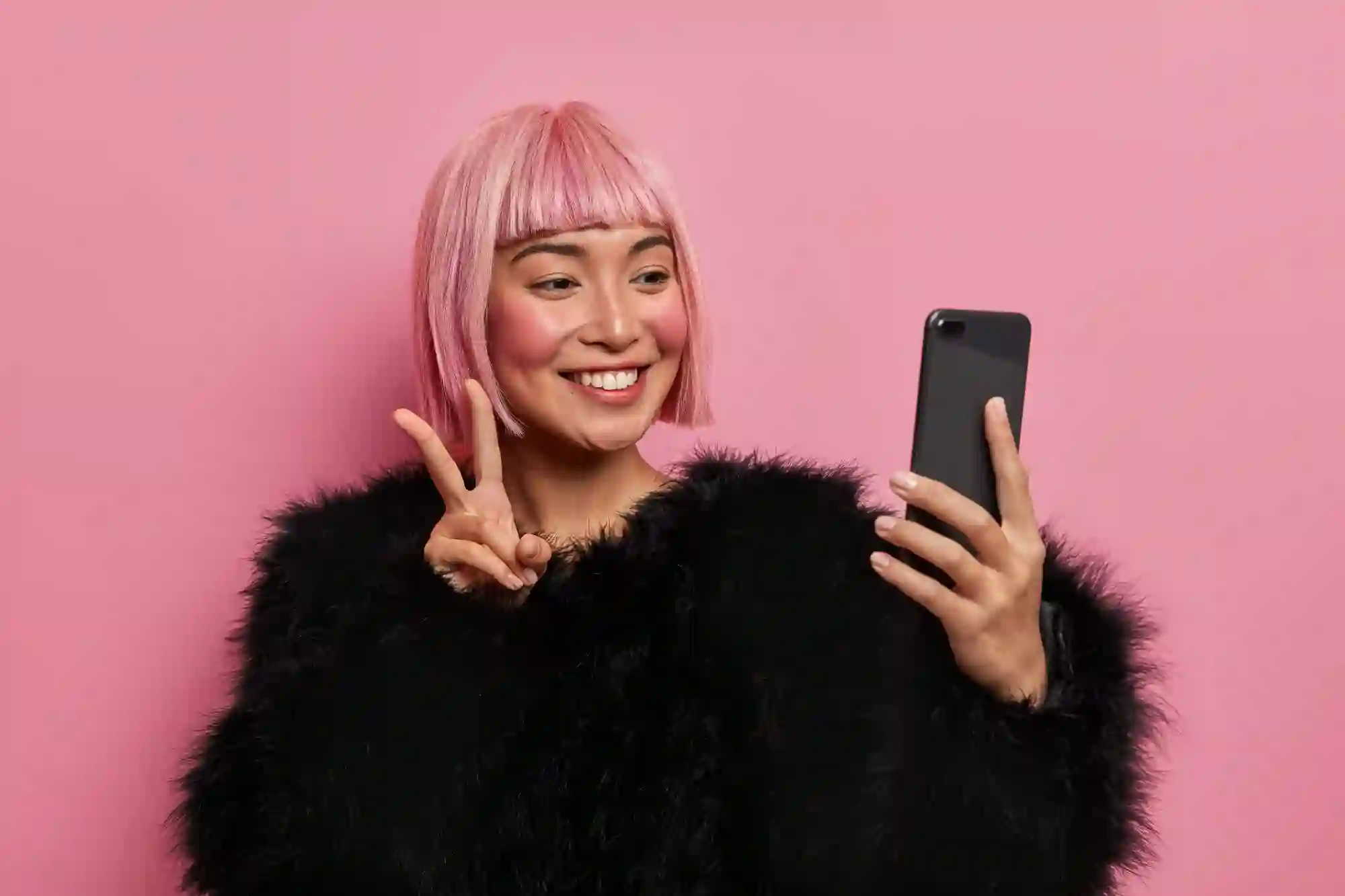 Smiling woman with pink hair and a black fluffy coat taking a selfie while flashing a peace sign.