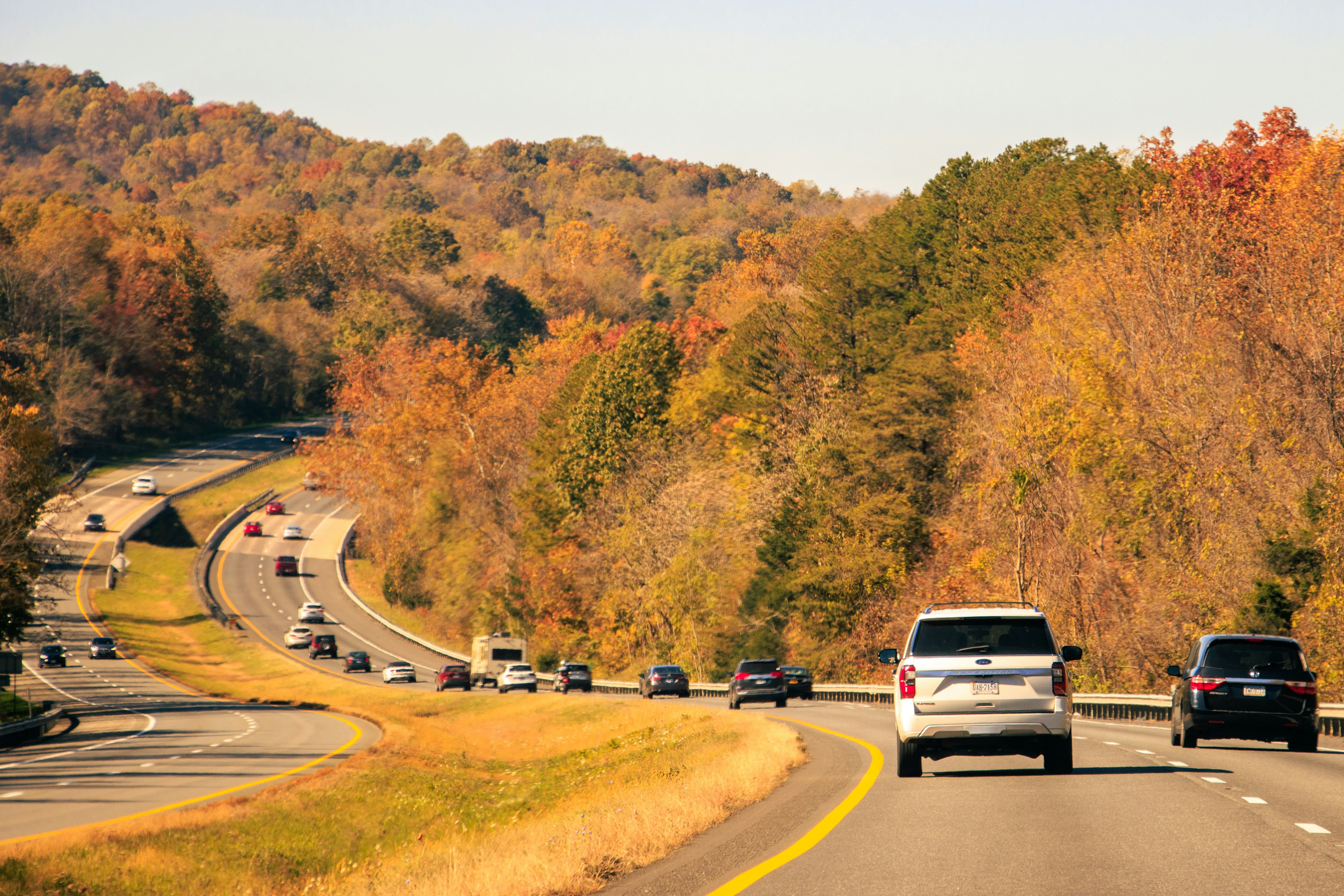 A scenic highway in Virginia, winding through forested hills during autumn. Cars travel along the road, surrounded by vibrant foliage in shades of green, yellow, and orange. The picturesque Virginia landscape under a clear sky highlights the beauty of the state’s countryside in fall.