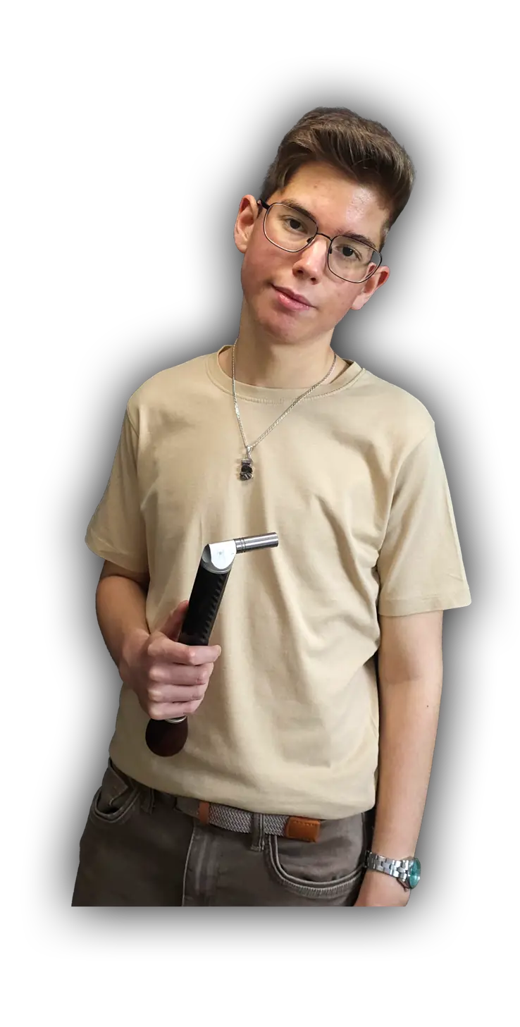 Adin Rrahmani, ein junger Mann mit kurzen braunen Haaren und rechteckiger Drahtbrille, steht vor einem weißen Hintergrund. Er trägt ein beiges, locker geschnittenes T-Shirt mit Rundhalsausschnitt, graue Jeans mit geflochtenem Gürtel und eine silberne Halskette mit kleinem Anhänger. An seinem rechten Handgelenk trägt er eine silberne Uhr mit türkisfarbenem Zifferblatt. In seiner linken Hand hält er aufrecht einen Klavierstimmhammer mit carbonummanteltem Griff und silbernem Metallkopf, leicht zur Kamera geneigt. Sein Blick ist ruhig und direkt, sein Ausdruck entspannt und gefasst.