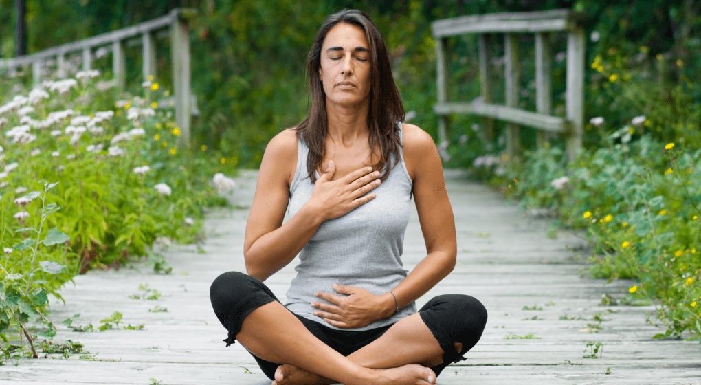An image of a woman sitting on the ground cross legged with her eyes closed, right hand over her chest and left hand on her stomach, with grass and a bridge in the background
