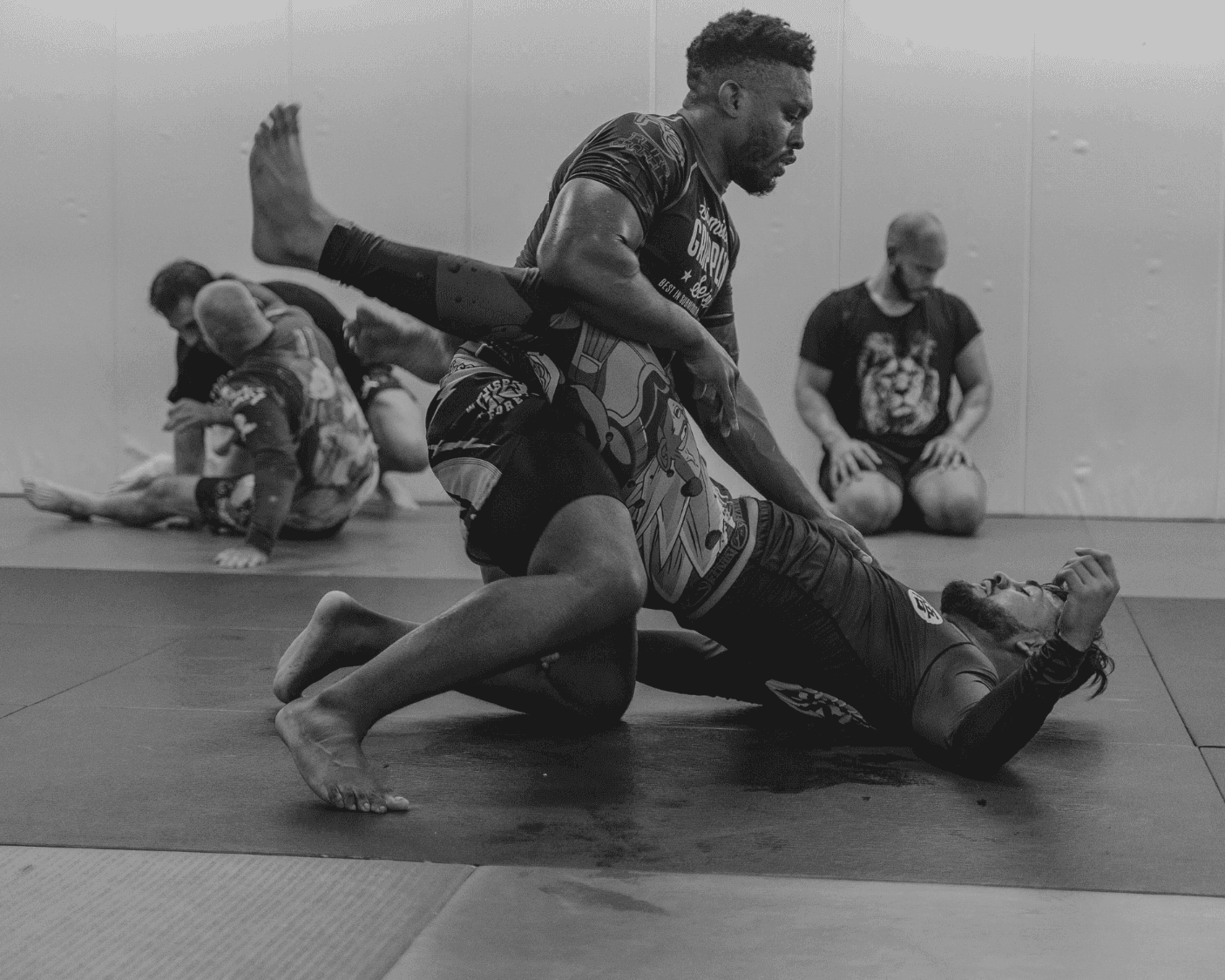 Two athletes engaged in intense no-gi Brazilian Jiu-Jitsu grappling on gym mats, with one fighter controlling position from the top while teammates look on in the background.