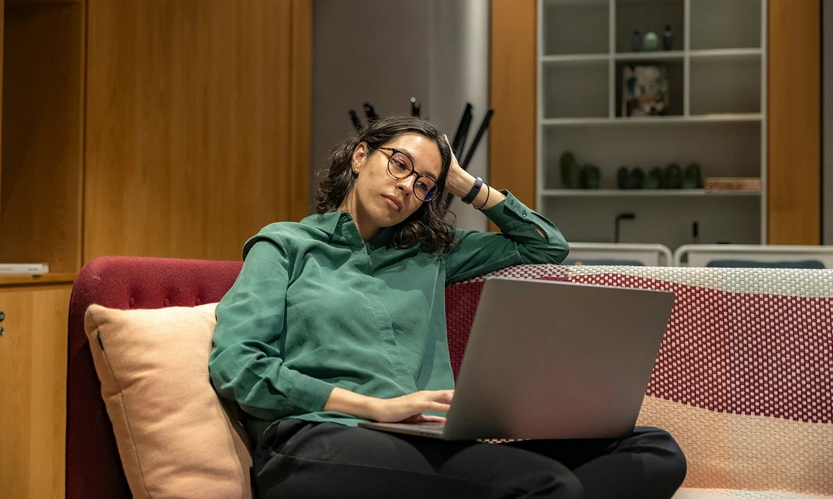 A person wearing glasses and a green shirt is sitting on a couch, holding their head with one hand while using a laptop, appearing thoughtful and relaxed in a modern, softly lit living room.