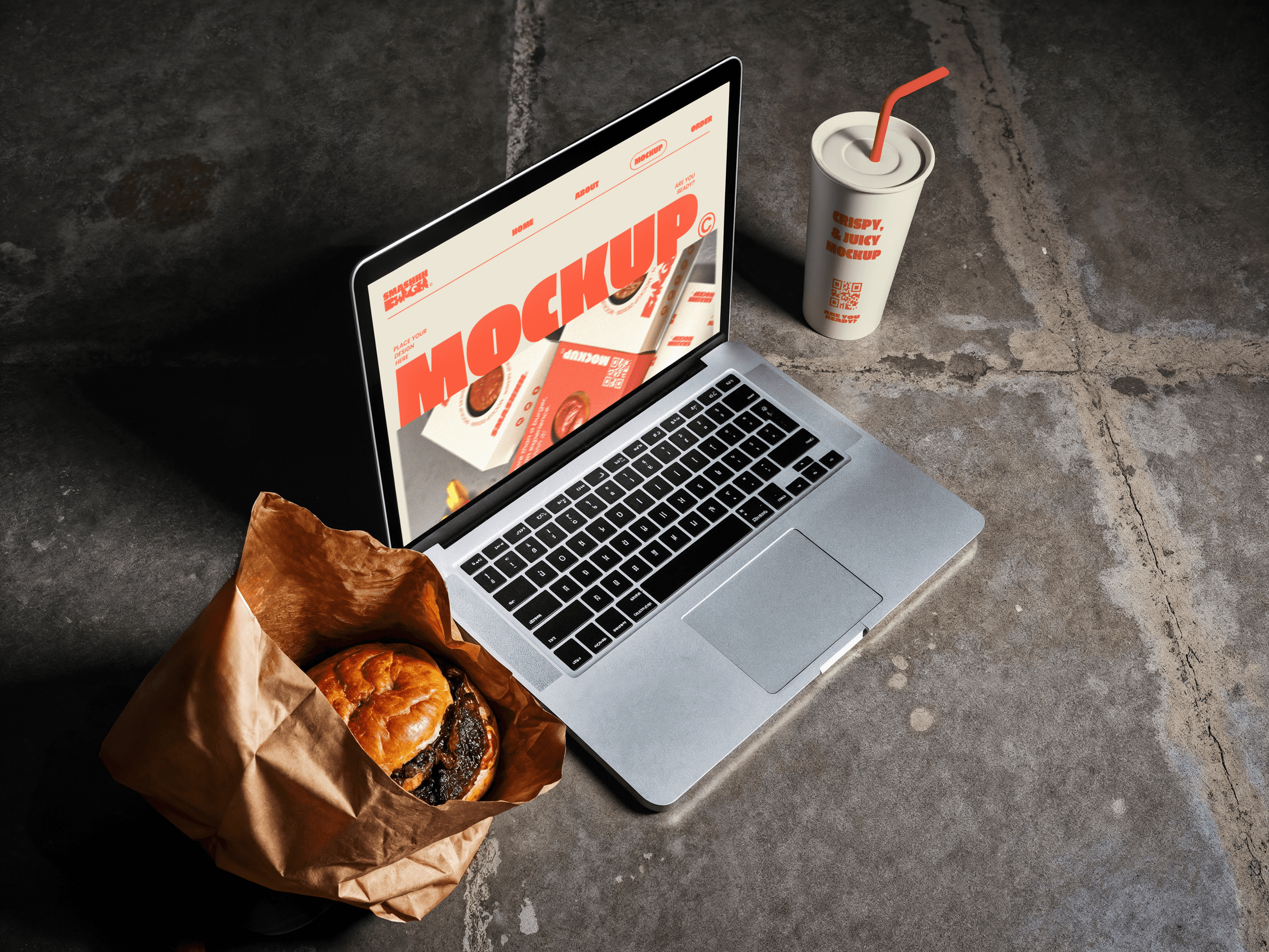 A top-down lifestyle mockup of an open laptop on a concrete surface next to a burger and a soda, displaying a bold "MOCKUP" design on the screen.