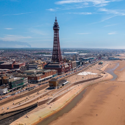 Beachfront cityscape with a tall tower, sandy beach, promenade, and scattered people under a clear blue sky.