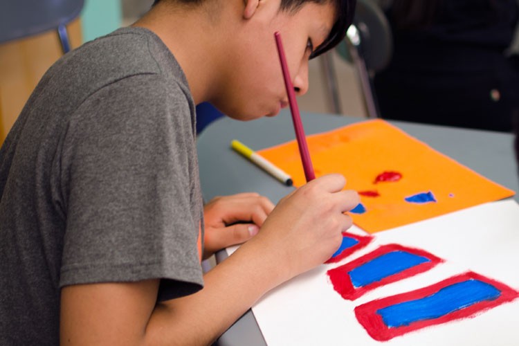 A young person with short dark hair, wearing a gray t-shirt, is seated at a table and painting a design with red and blue paint on a white sheet of paper. The person is using a red paintbrush in their right hand and a yellow marker in their left hand. A second piece of orange paper with blue and red paint remnants rests on the table near the painting. A portion of a blue chair is visible in the background.