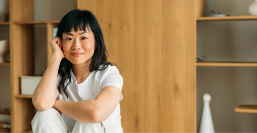 A woman named Jenny O with black hair smiles while sitting on a wooden surface, with shelves in the background.