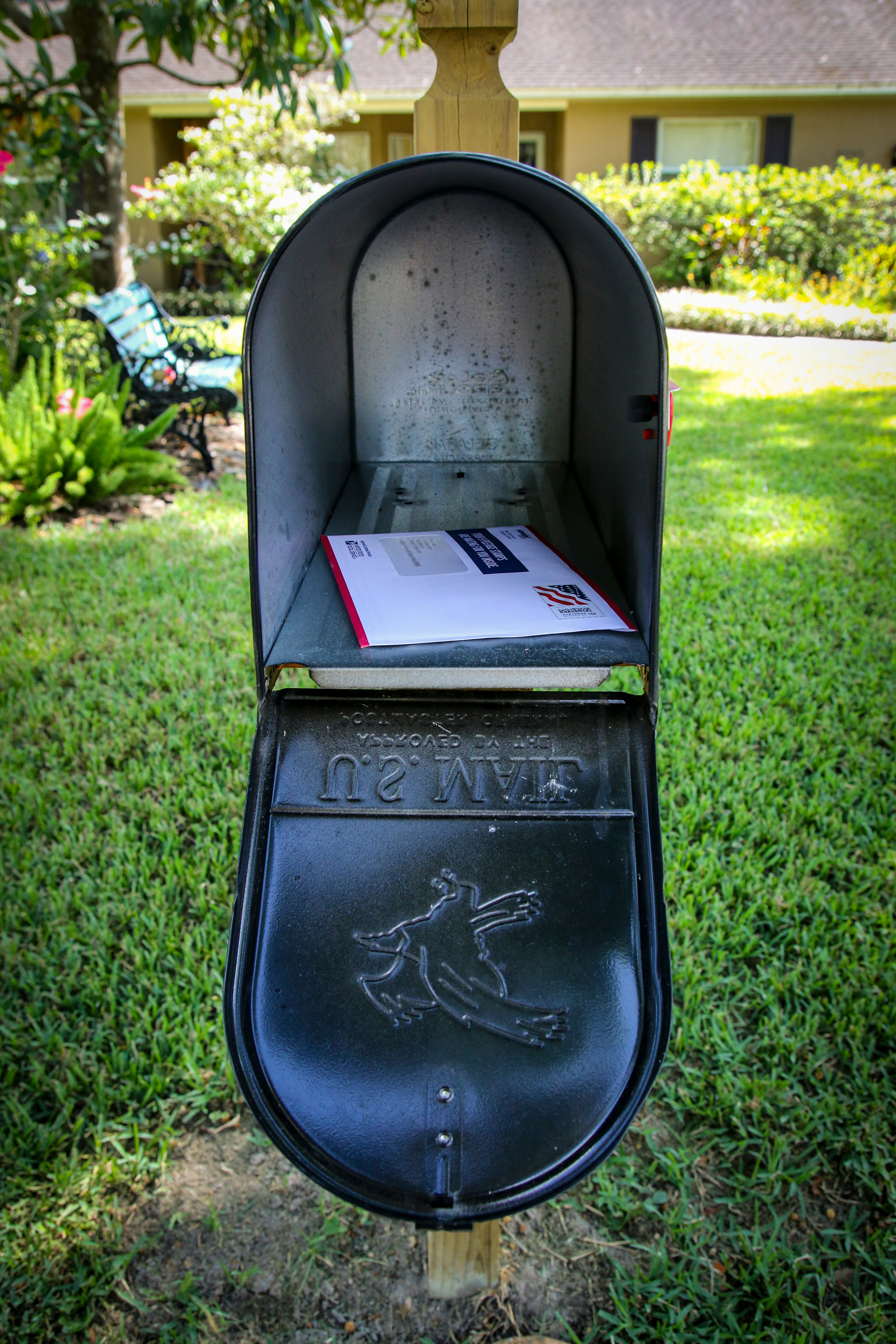Open residential mailbox with an official government envelope inside, set against a suburban home and lawn.
