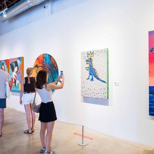 Persone che osservano opere d'arte colorate su una parete della galleria; una donna scatta una foto con il suo telefono.