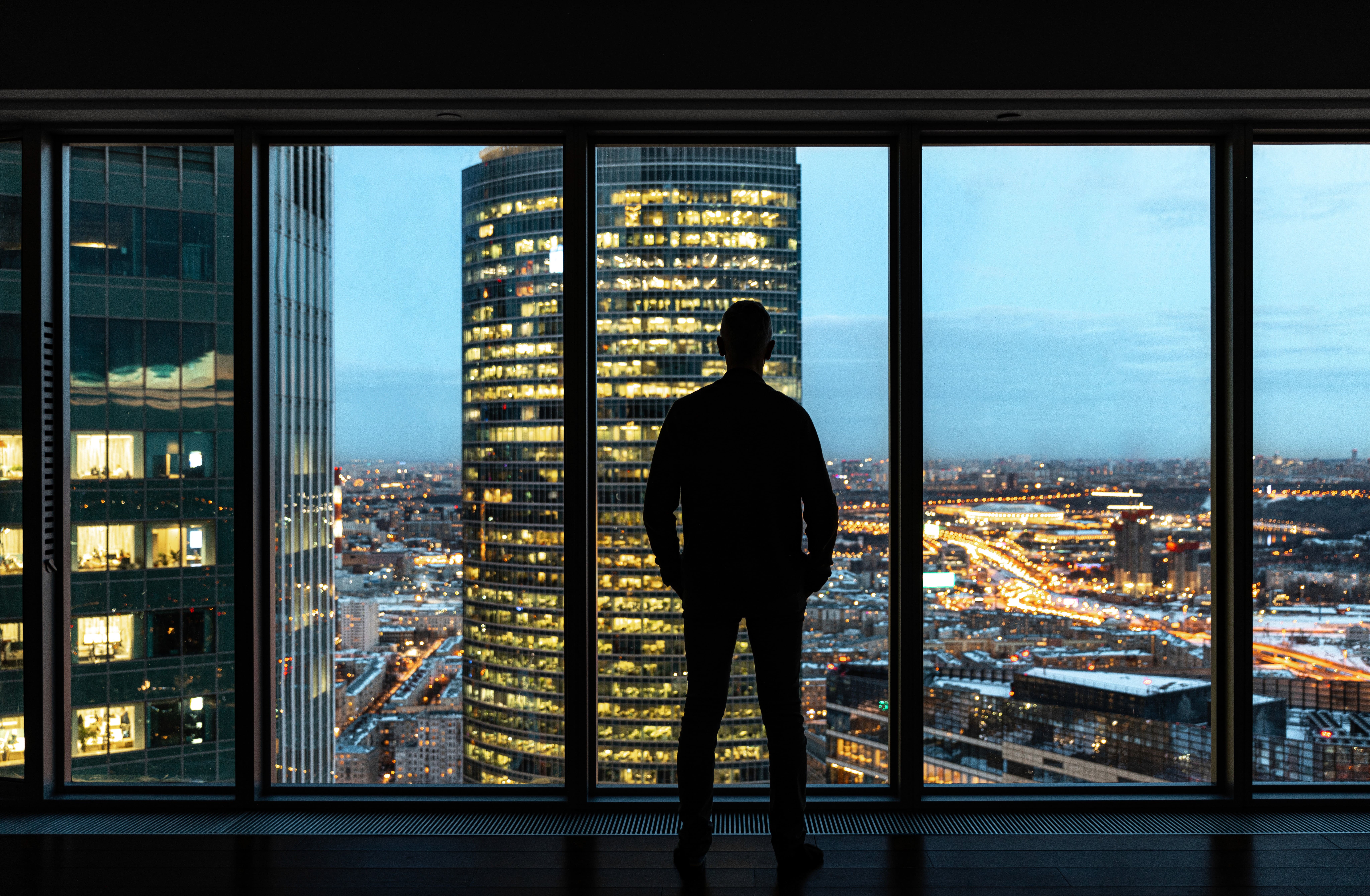 Silhouette of a person at a window, overlooking a lit city skyline at night.