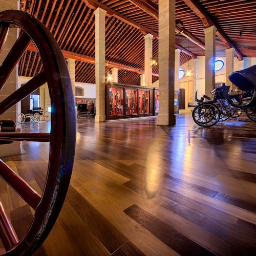 Spacious museum hall with wooden floors, display cases, vintage carriages, and a red wagon wheel in the foreground.