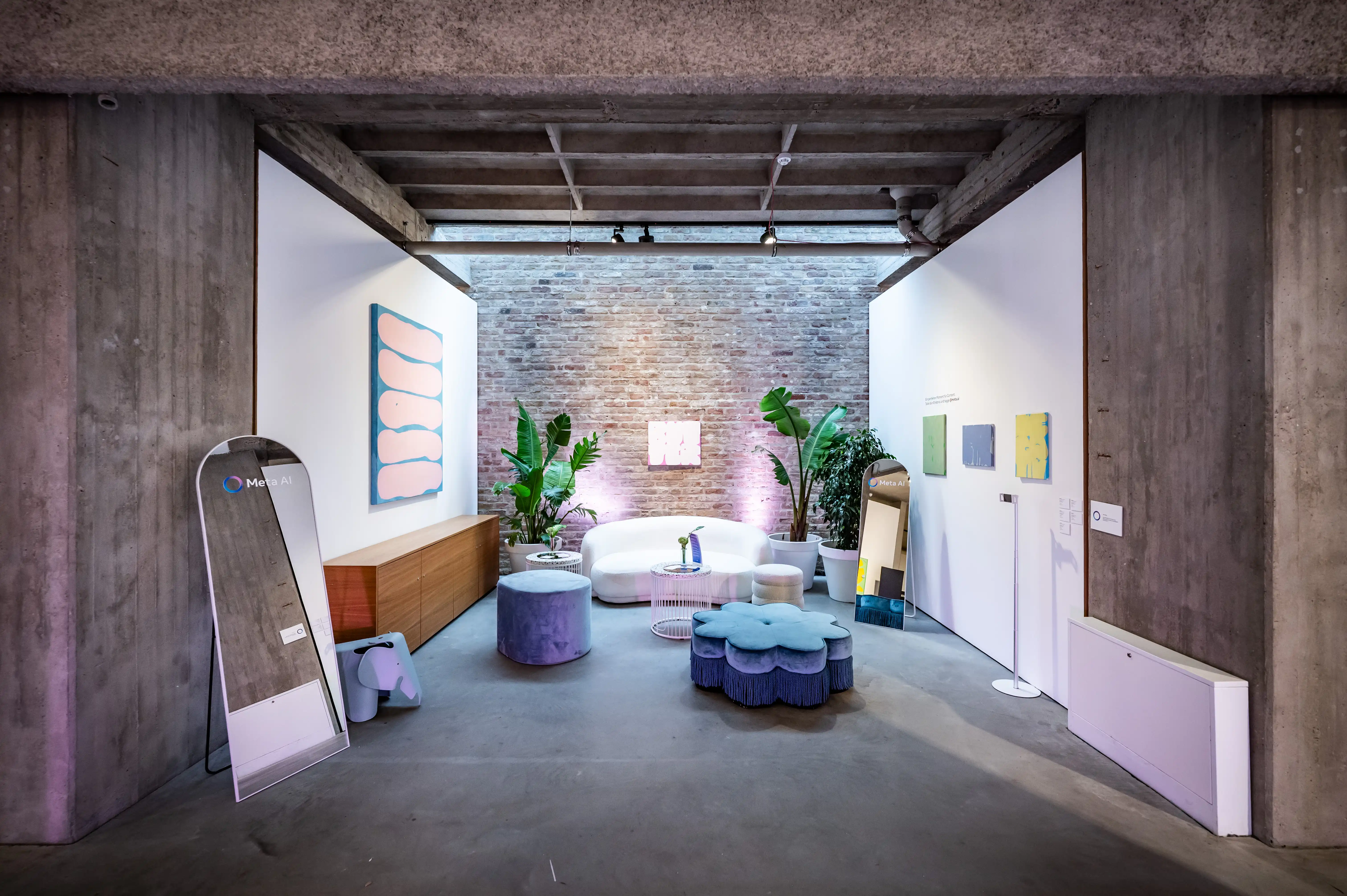 Showroom with white sofa, lilac poufs, plants and pastel artworks between concrete pillars.