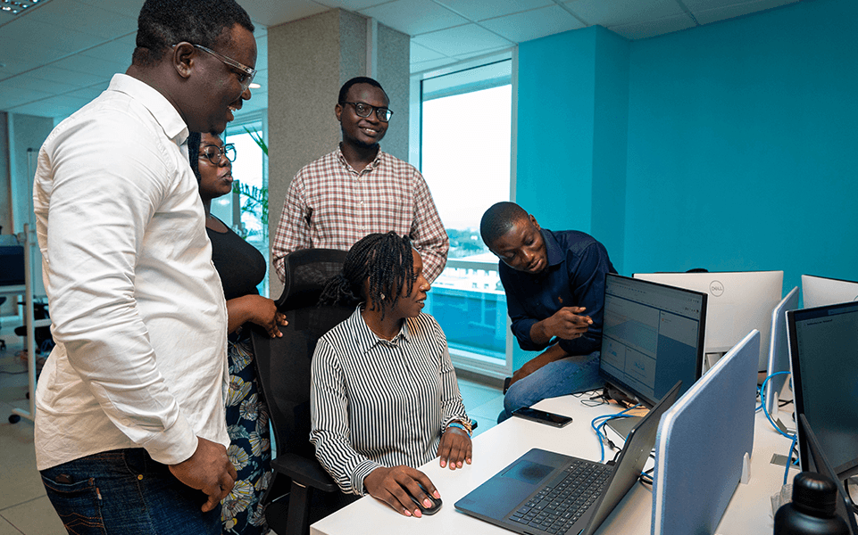 A group of young professionals gathered around a laptop, discussing and collaborating in a modern office setting.