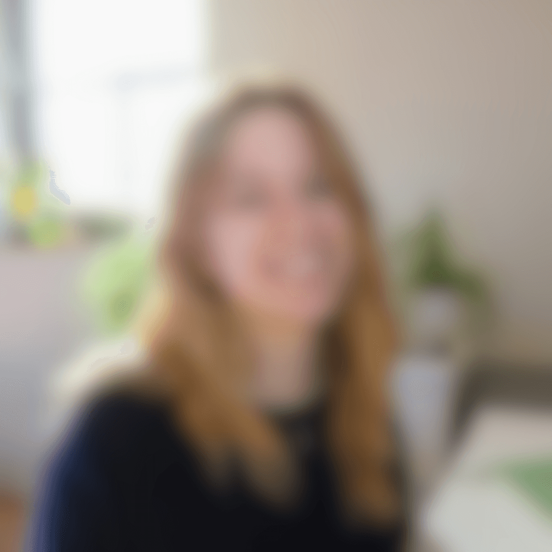Zwei Personen sitzen an einem hellen Schreibtisch in einem Coworking Space. Die Person im Vordergrund trägt einen cremefarbenen Pullover und weite Jeans, hat lange rotbraune Haare und hält ein Smartphone in der Hand. Auf dem Tisch stehen ein Laptop, ein Glas Wasser und eine Vase mit hellen Tulpen. Die Umgebung wirkt modern und minimalistisch, mit neutralen Wandfarben und einem dunklen Boden.