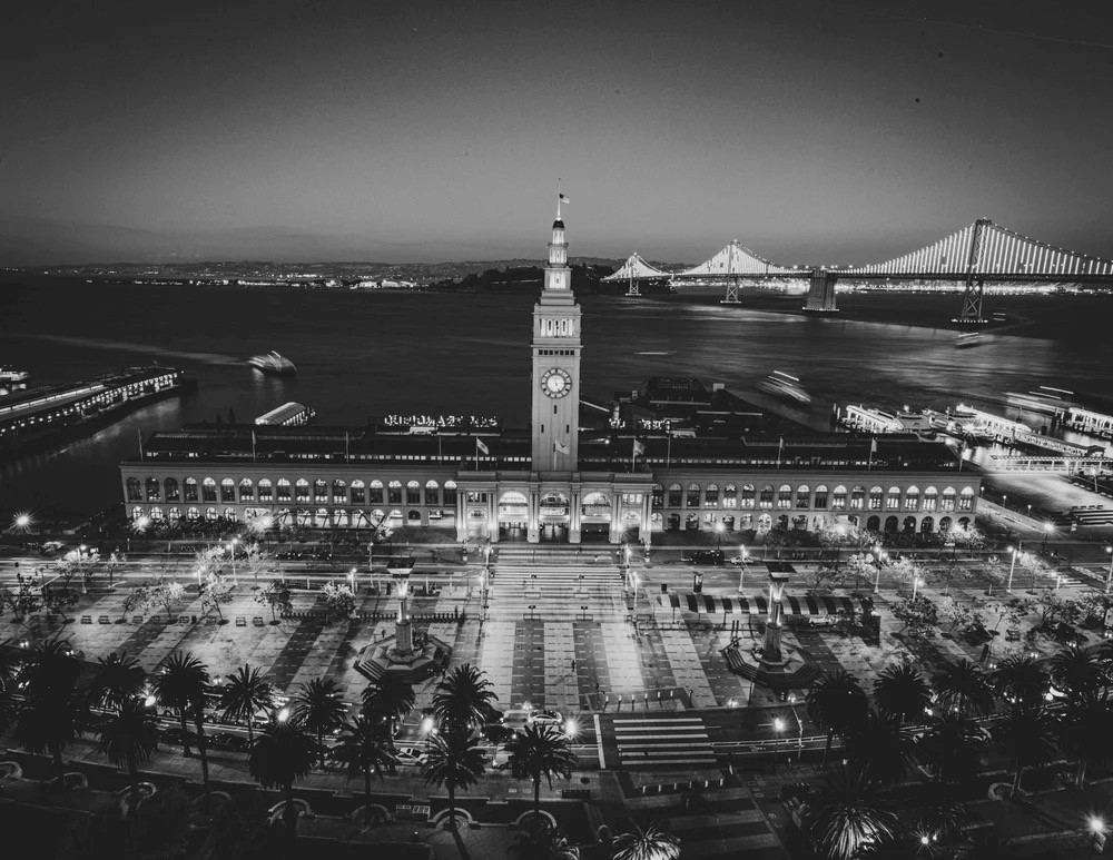 Black and white aerial view of SF Embarcadero building with lit up Bay Bridge in background