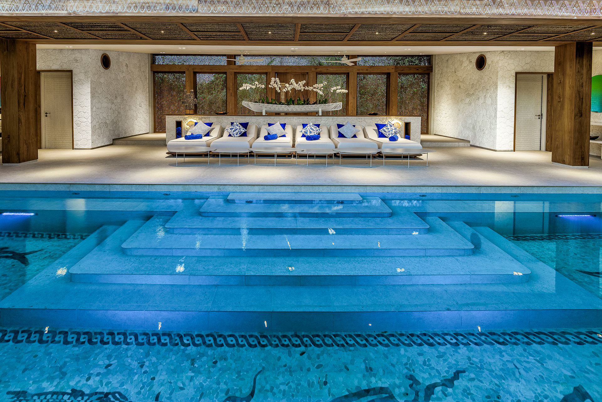 Innenansicht eines luxuriösen Wellnessbereichs mit einer Reihe von sechs weißen Sonnenliegen mit blauen Kissen, die vor einer kunstvoll geschnitzten Holzwand an einem blau beleuchteten Indoor-Pool stehen.