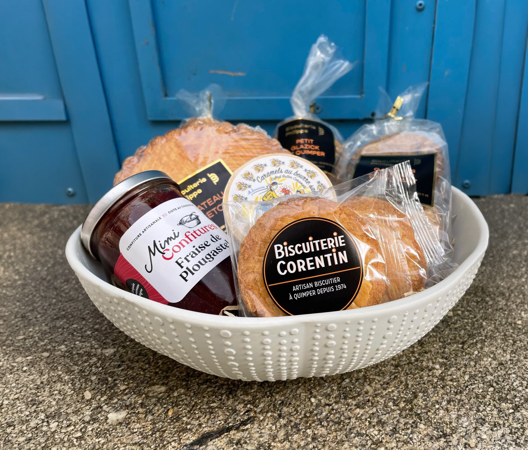Bol en céramique blanche à relief de pointillés présentant un panier gourmand avec gâteau breton, confiture et biscuits, devant une porte bleue