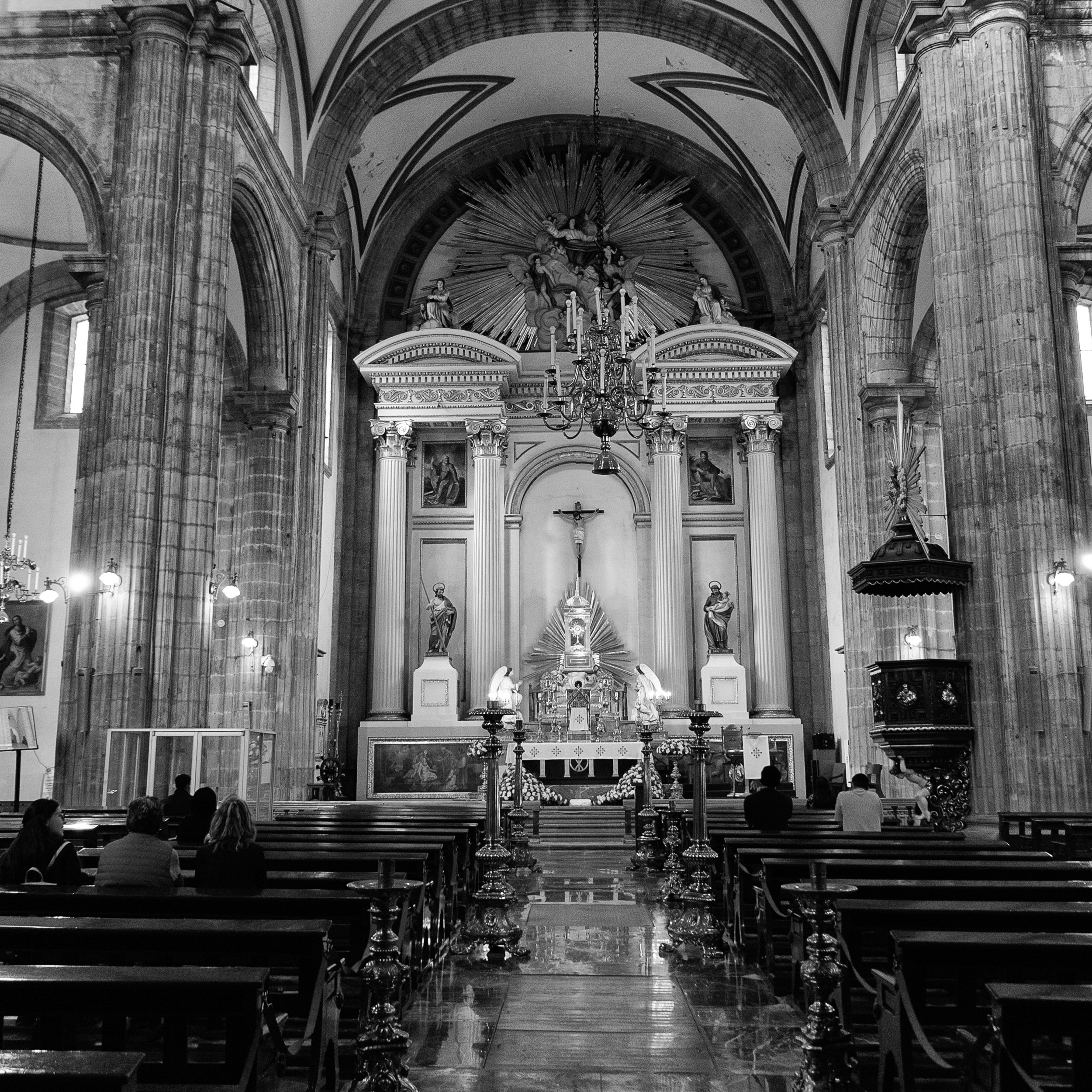 Black and white photograph of the interior of Mexico City's cathedral, showing the ornate altar, tall stone columns, and people seated in quiet prayer.