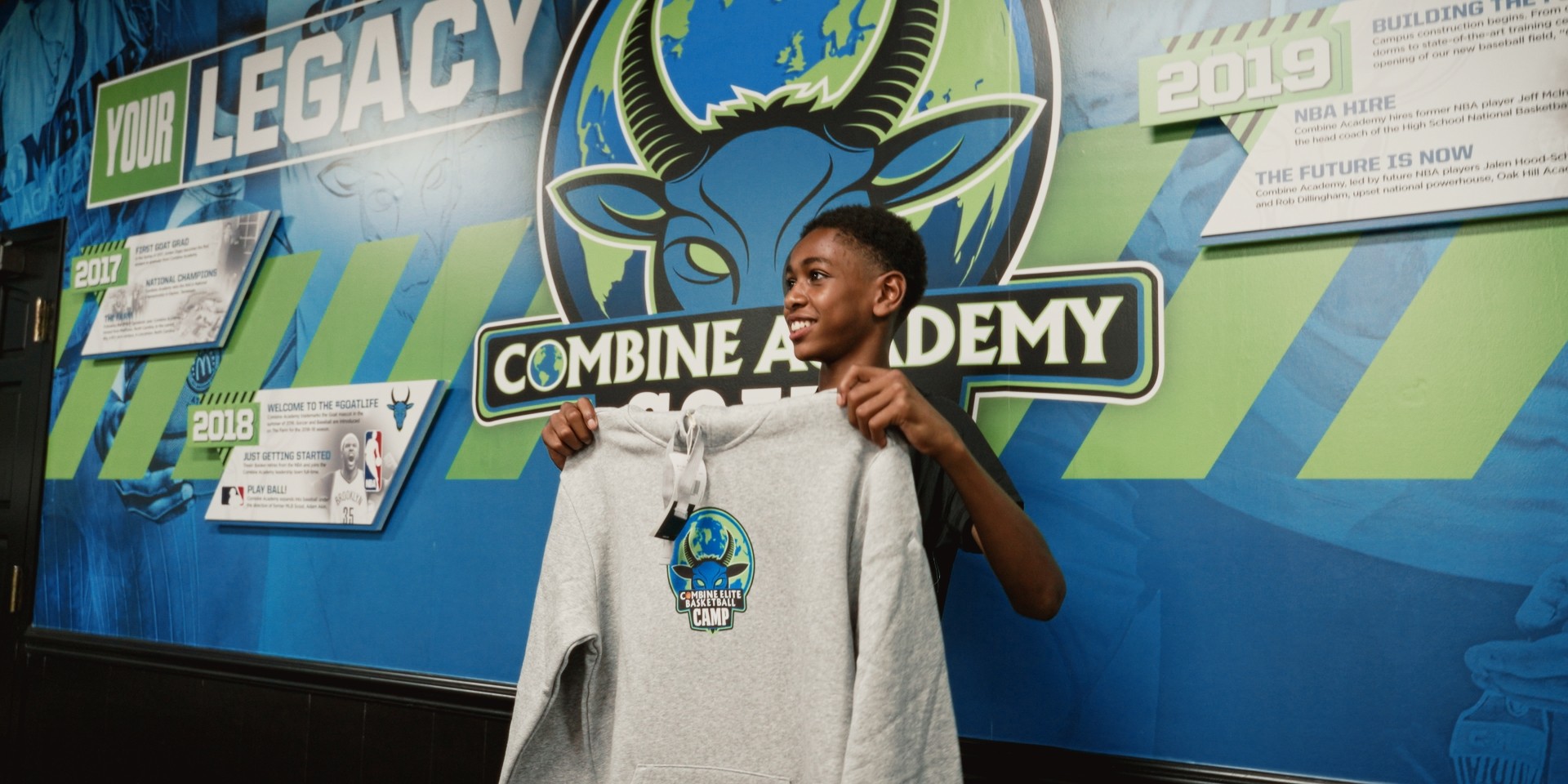 Jeune participant souriant tenant un sweat du Combine Elite Camp devant un mur décoré.