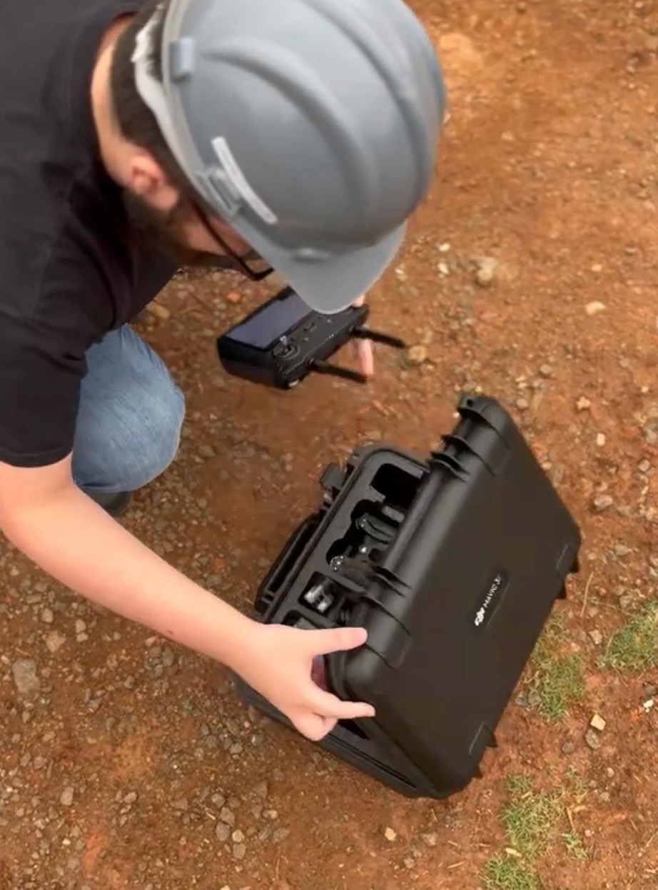 Um colaborador com capacete cinza agachado no canteiro de obras, abrindo uma maleta rígida de transporte reforçada que contém um drone e acessórios. Mostra o cuidado com o equipamento de Captura da Realidade no dia a dia da obra.